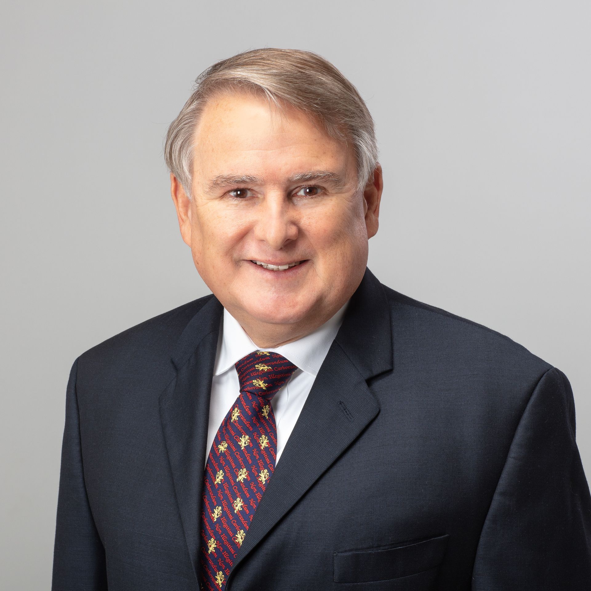 Man in dark suit, smiling, wearing patterned tie, against a gray background.