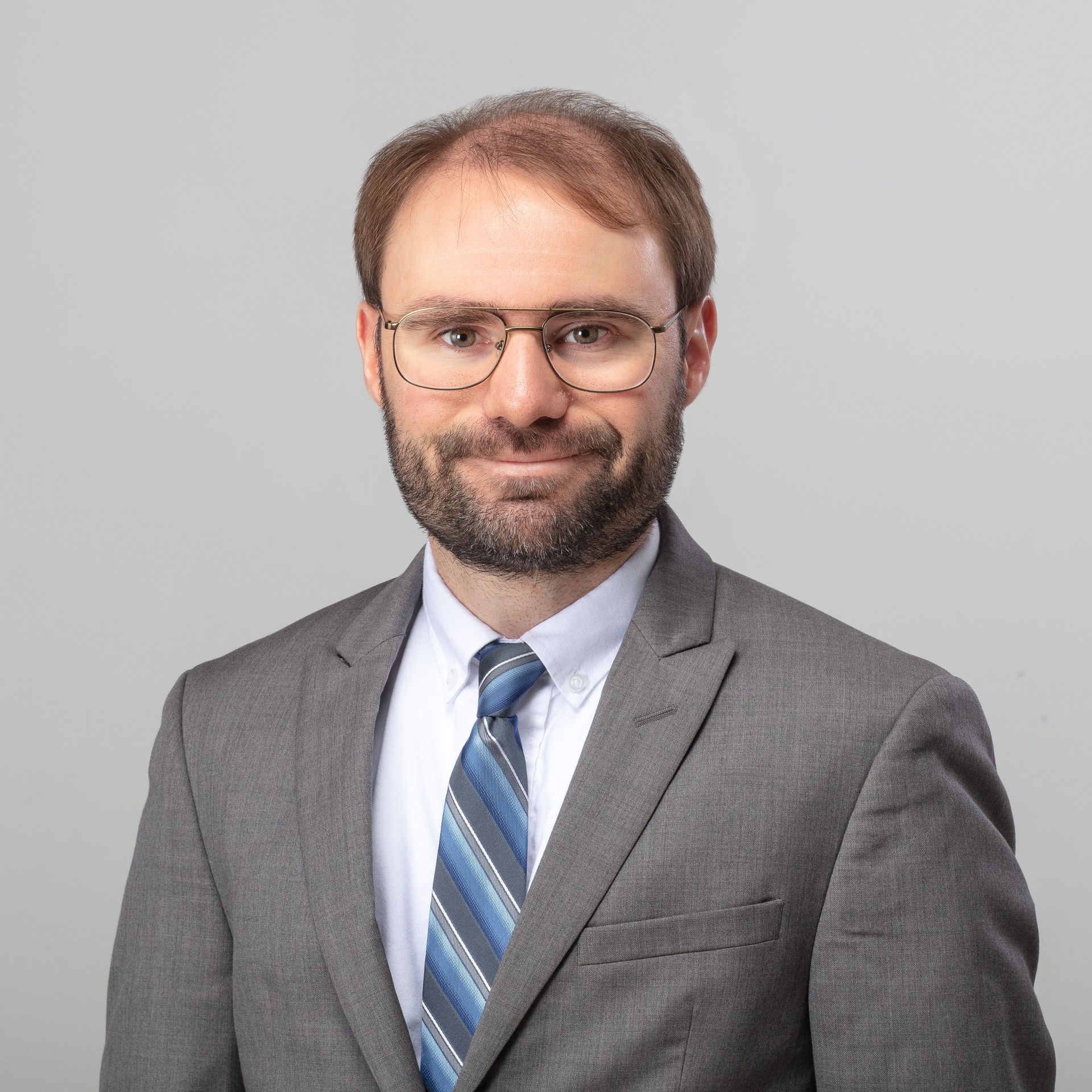 Man in glasses, gray suit, and blue striped tie smiles, studio shot with gray background.