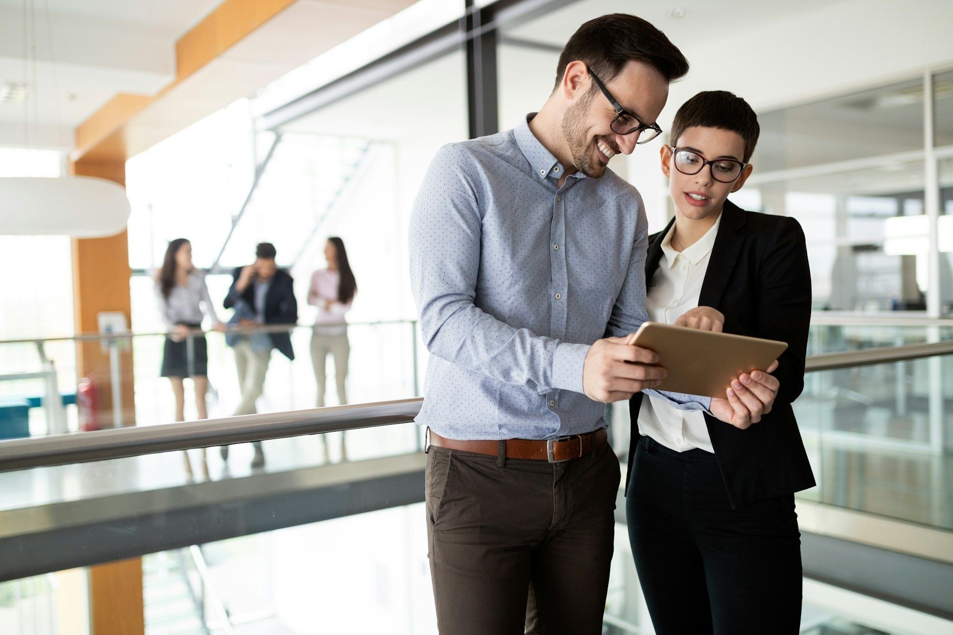 Two coworkers looking at tablet, smiling, in modern office setting with other people blurred in background.