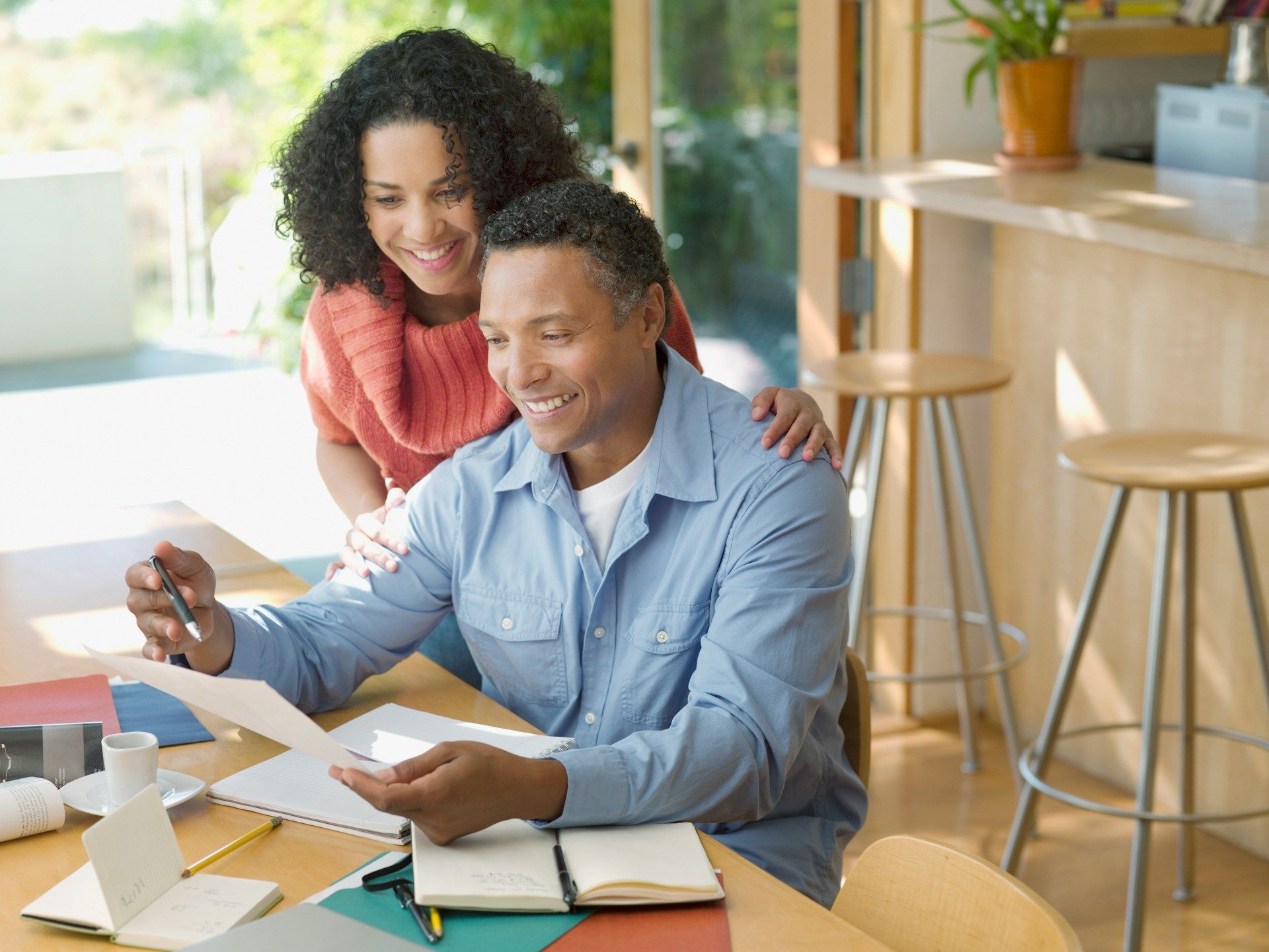 A couple reviews documents together at a table. Woman has her arm around man's shoulders, smiling.