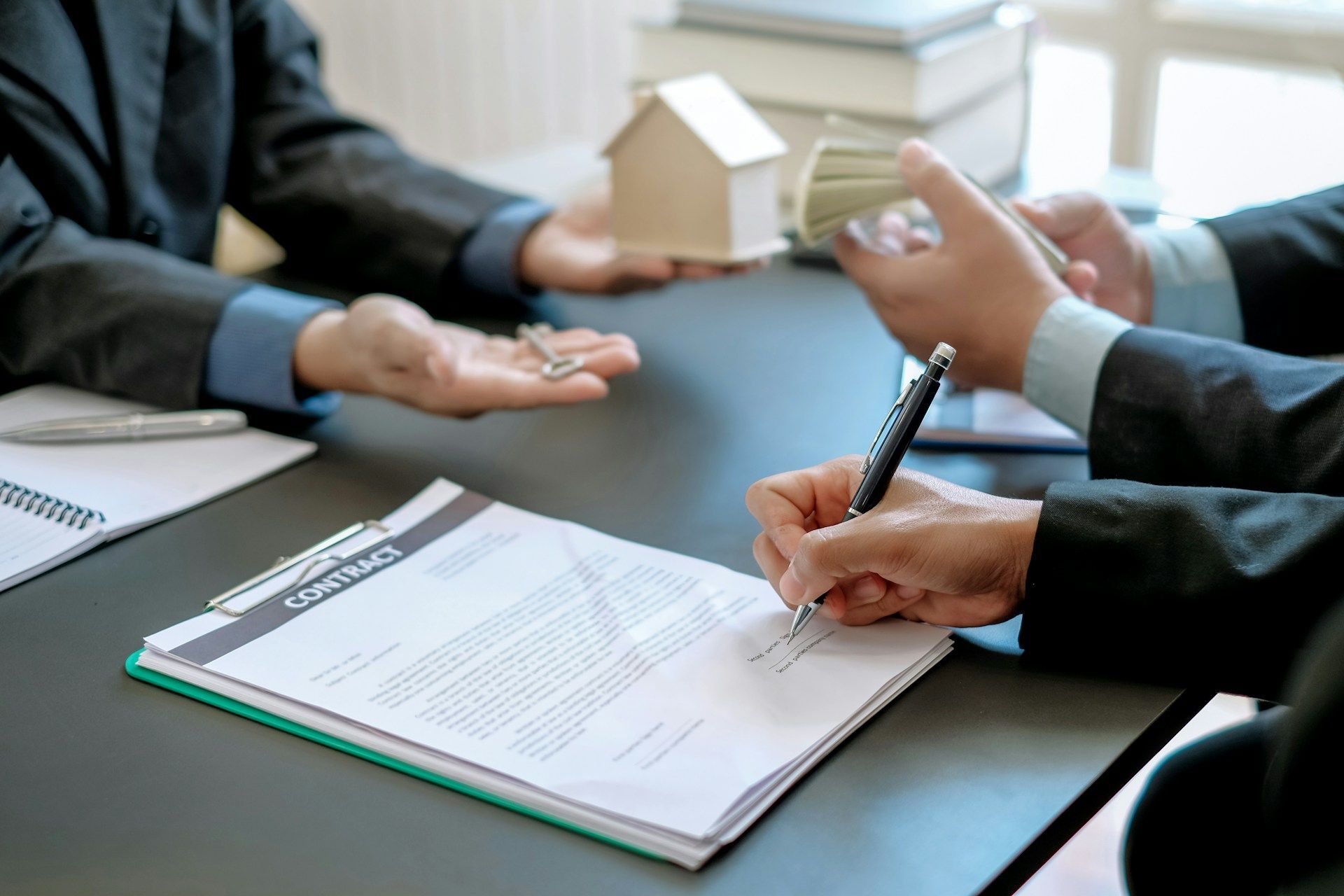 Person signing a contract with others, house model, keys, and money on a table.