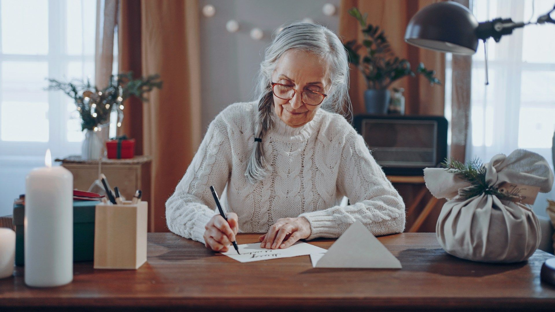 Woman writing at a desk indoors, in a festive setting with candles and decorations.