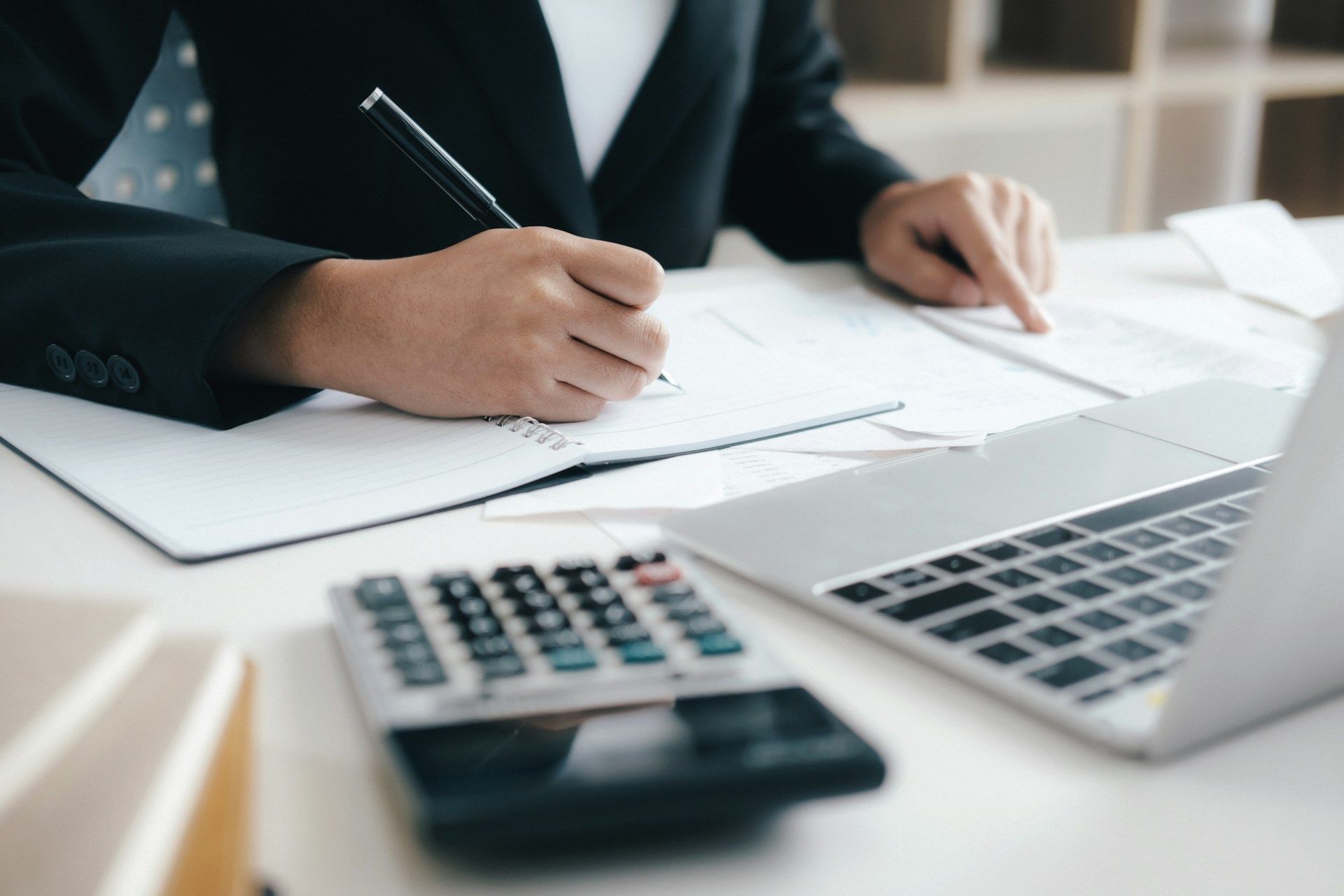 Person in suit writing in notebook next to laptop and calculator.