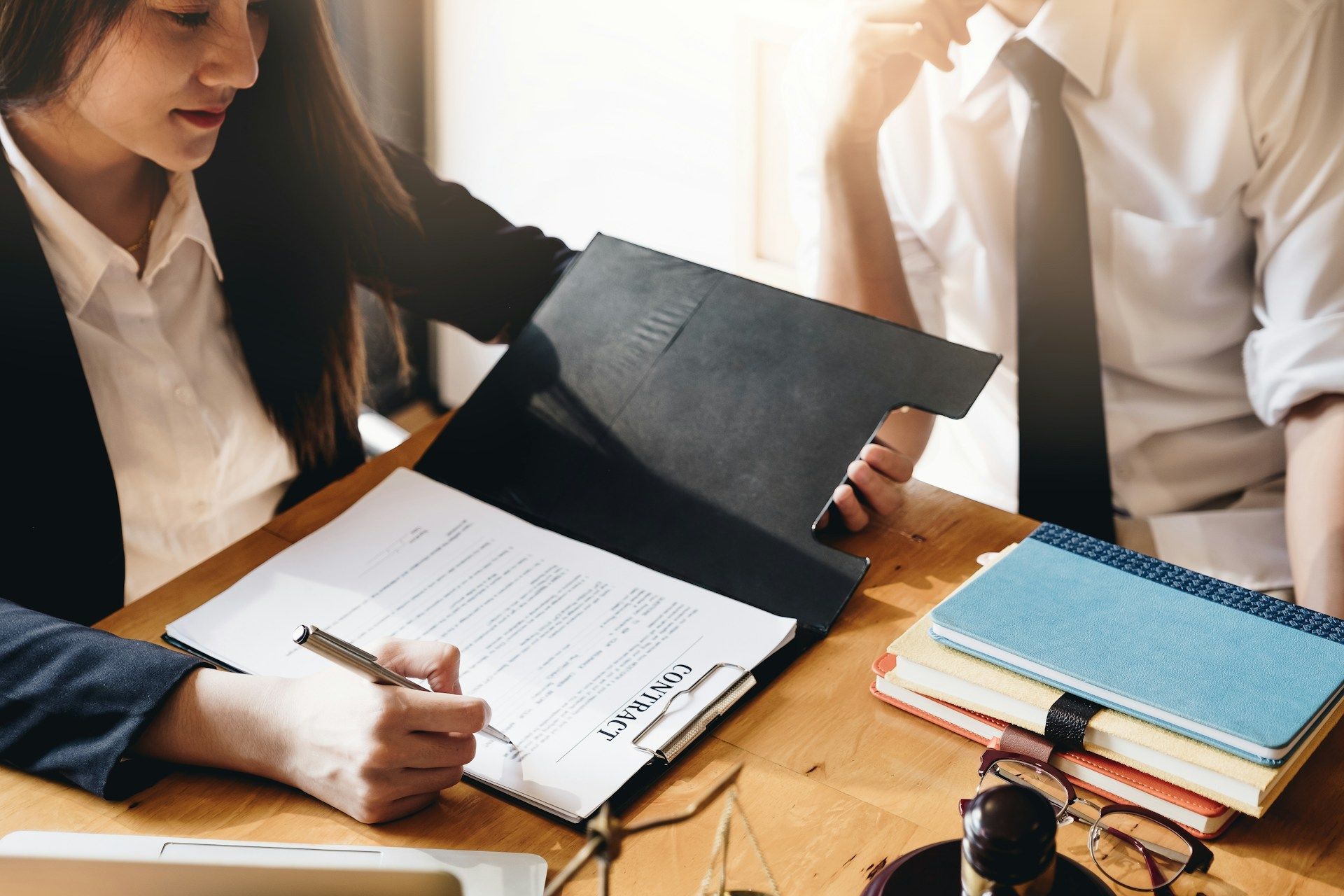 Woman signing a document in a folder on a wooden desk with a man in a tie looking on. Stack of books and eyeglasses nearby.