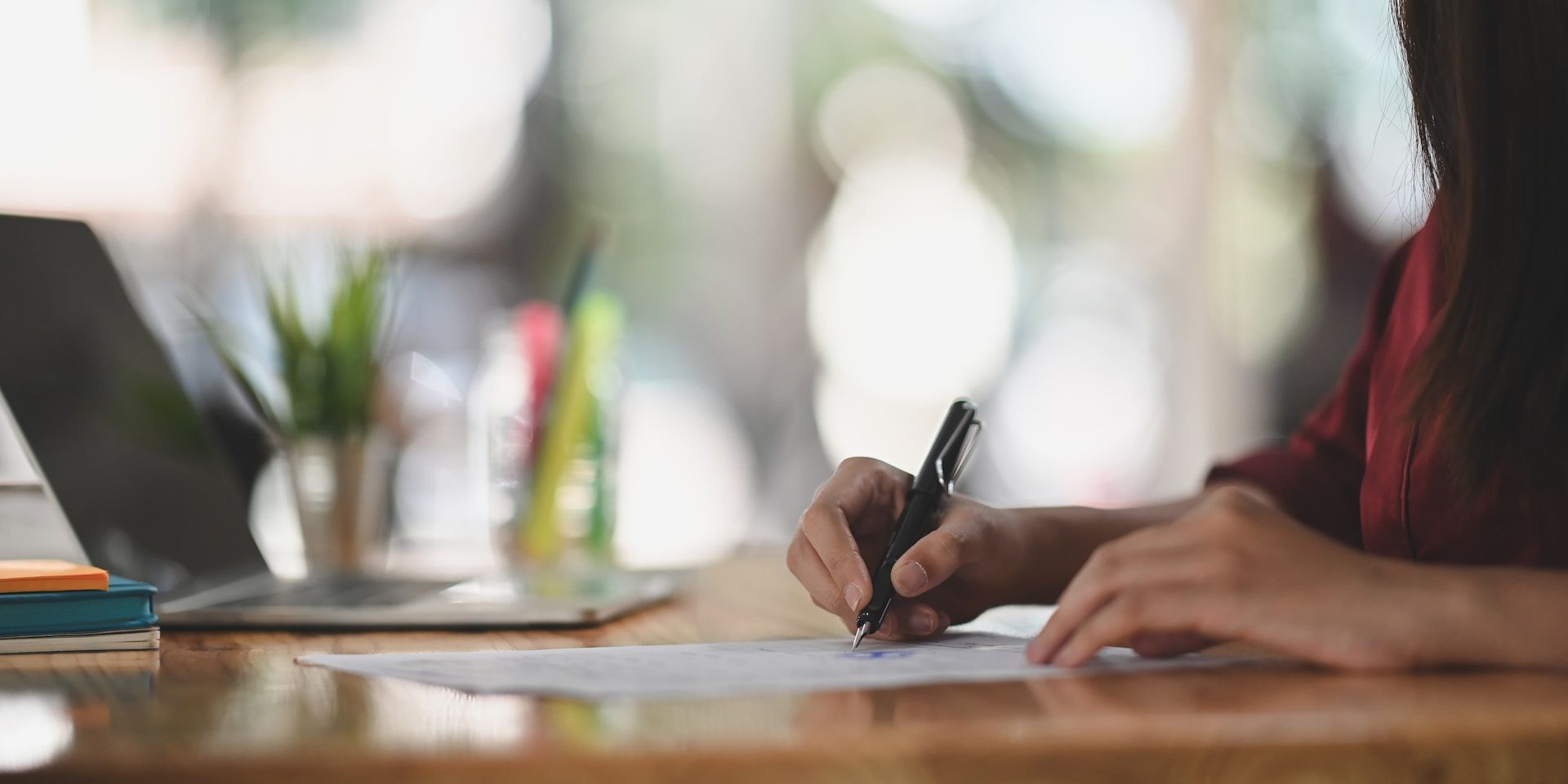 Person writing on paper at a wooden desk, near a laptop and other items. Bokeh background.