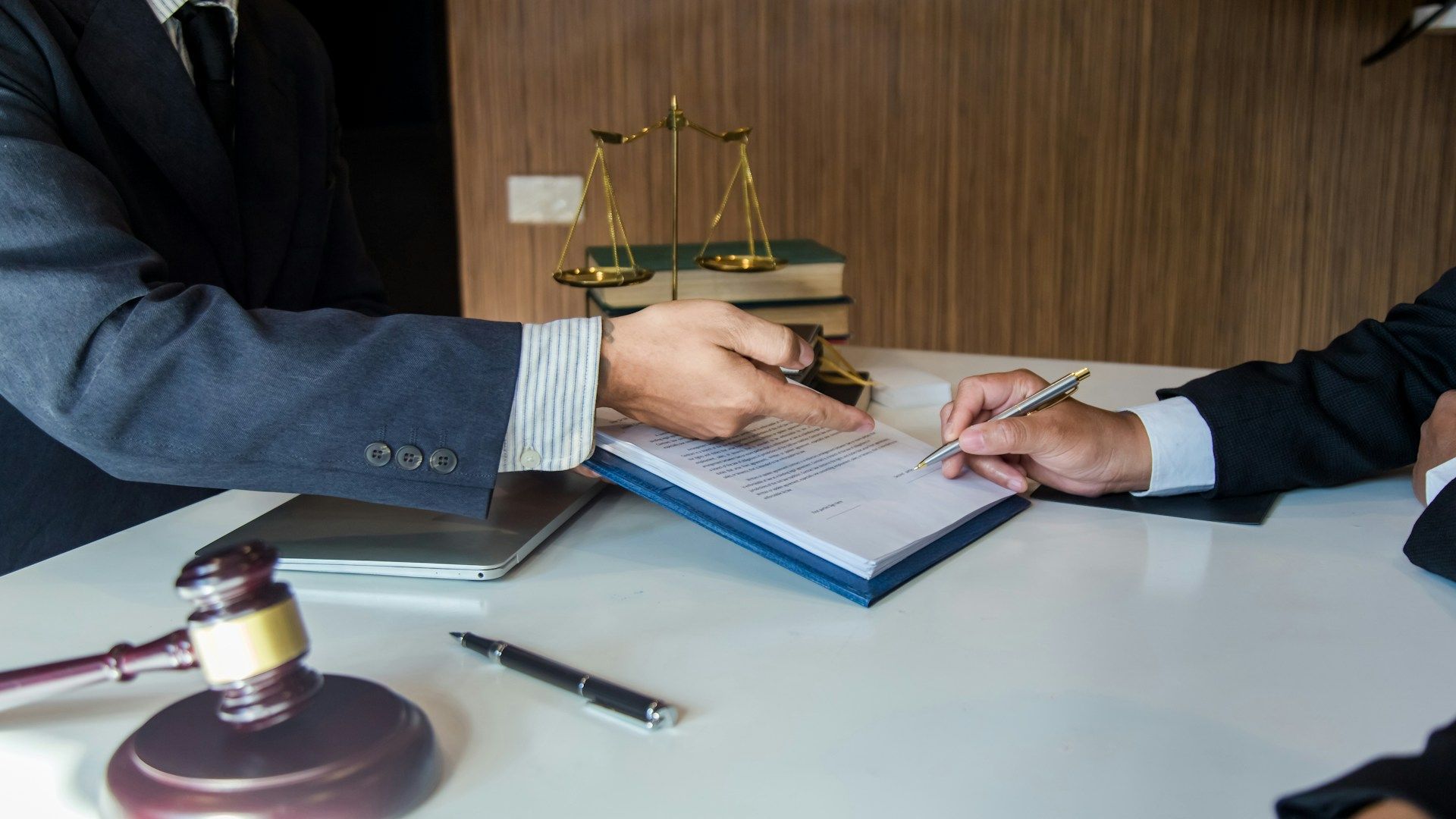 Two men in suits at a desk, one signing a document, scales of justice and gavel in the background.