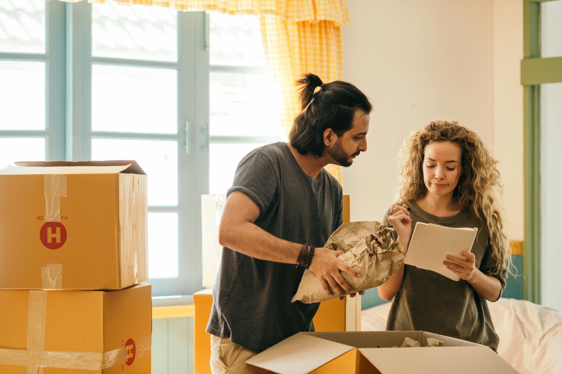 Couple packing boxes, one holding wrapped item, other looking at tablet, moving.