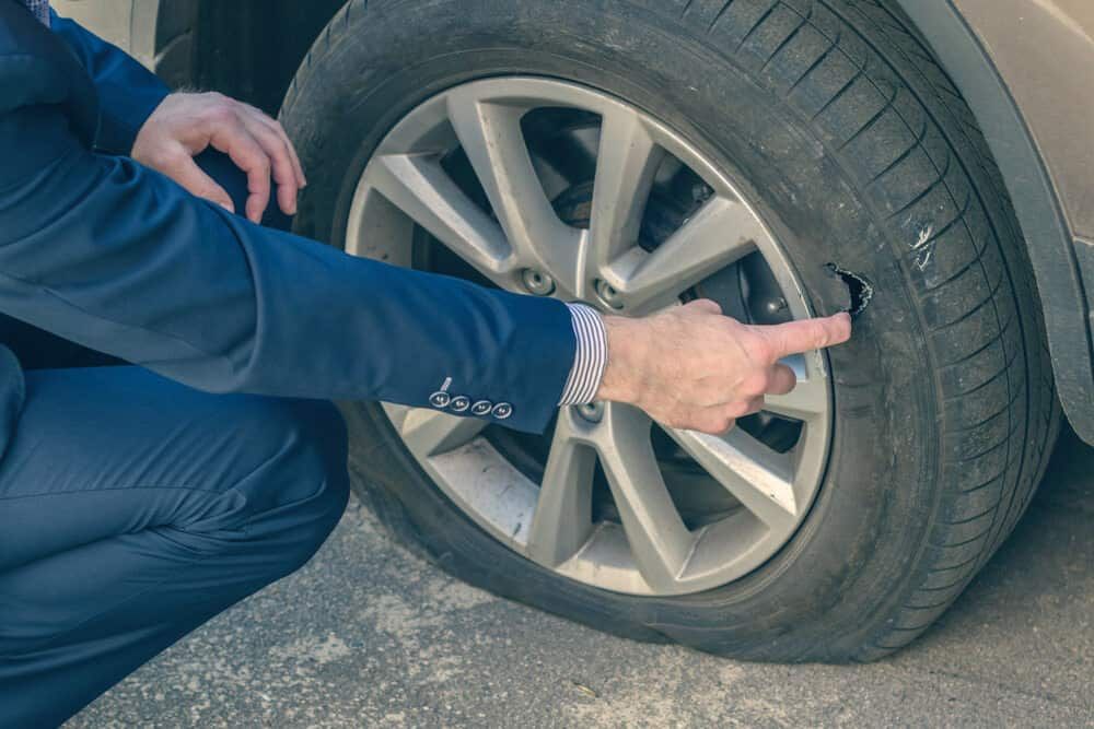 A Man In A Suit Is Pointing At A Tire That Has A Hole In It — Manning Smash & Auto Repairs In Wingham, NSW