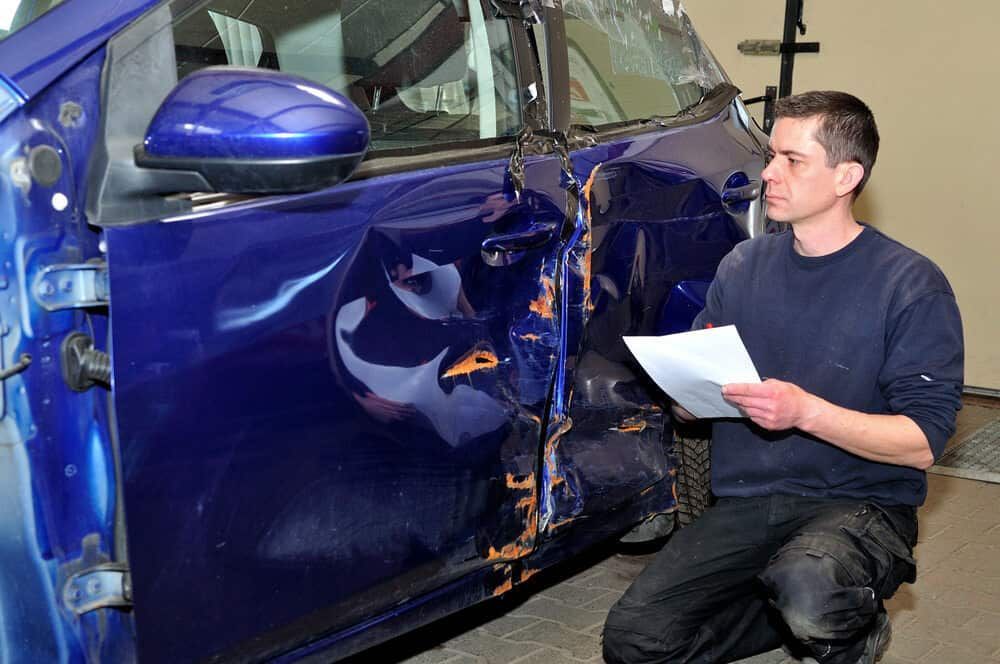 A Man Is Kneeling Down In Front Of A Blue Car That Has Been Damaged — Manning Smash & Auto Repairs In Wingham, NSW