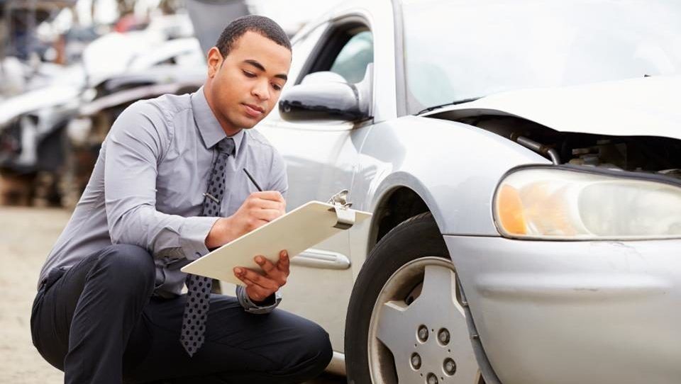 A Man Is Kneeling Down Next To A Car And Writing On A Clipboard — Manning Smash & Auto Repairs In Wingham, NSW