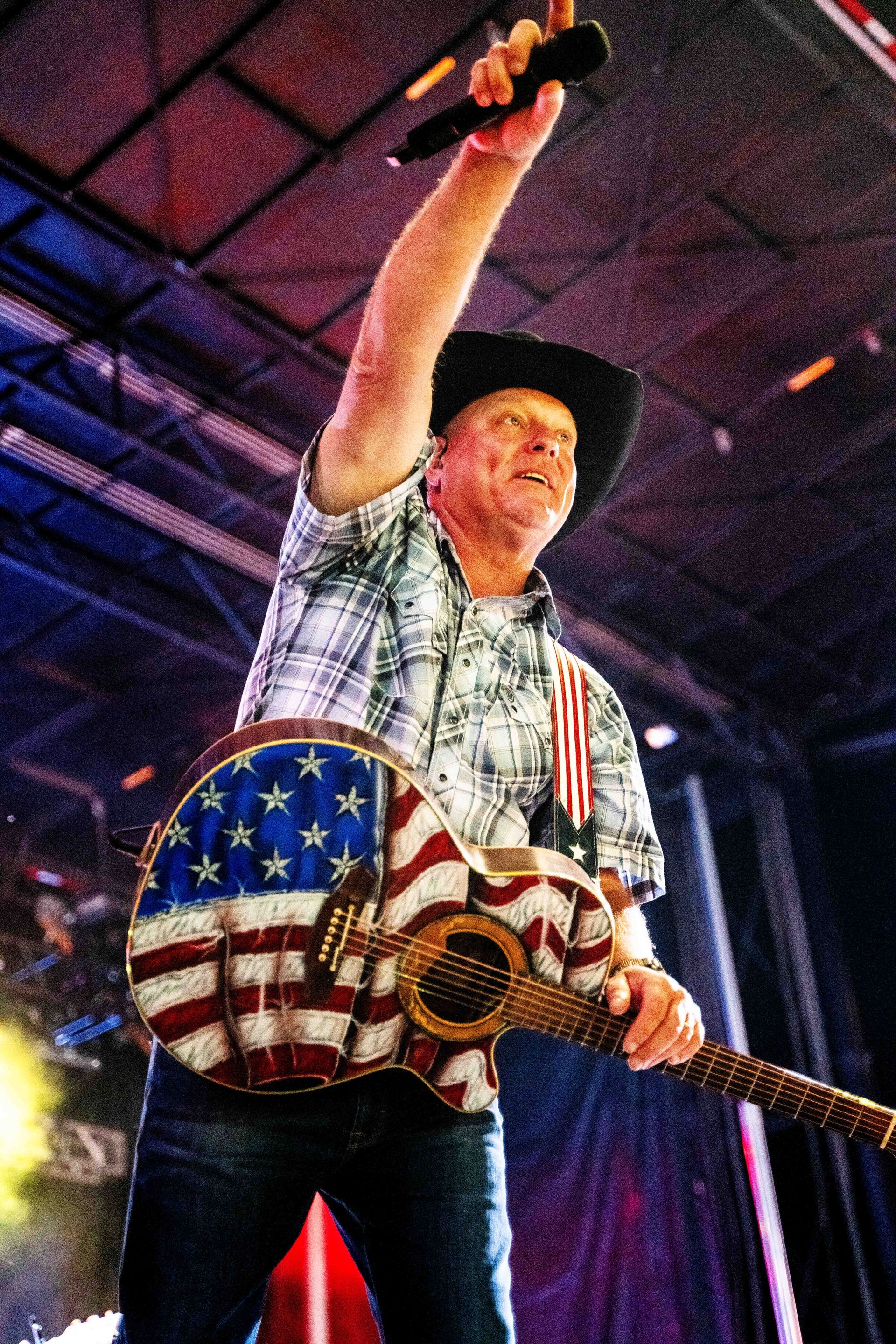 A man in a cowboy hat is singing into a microphone while holding an american flag guitar