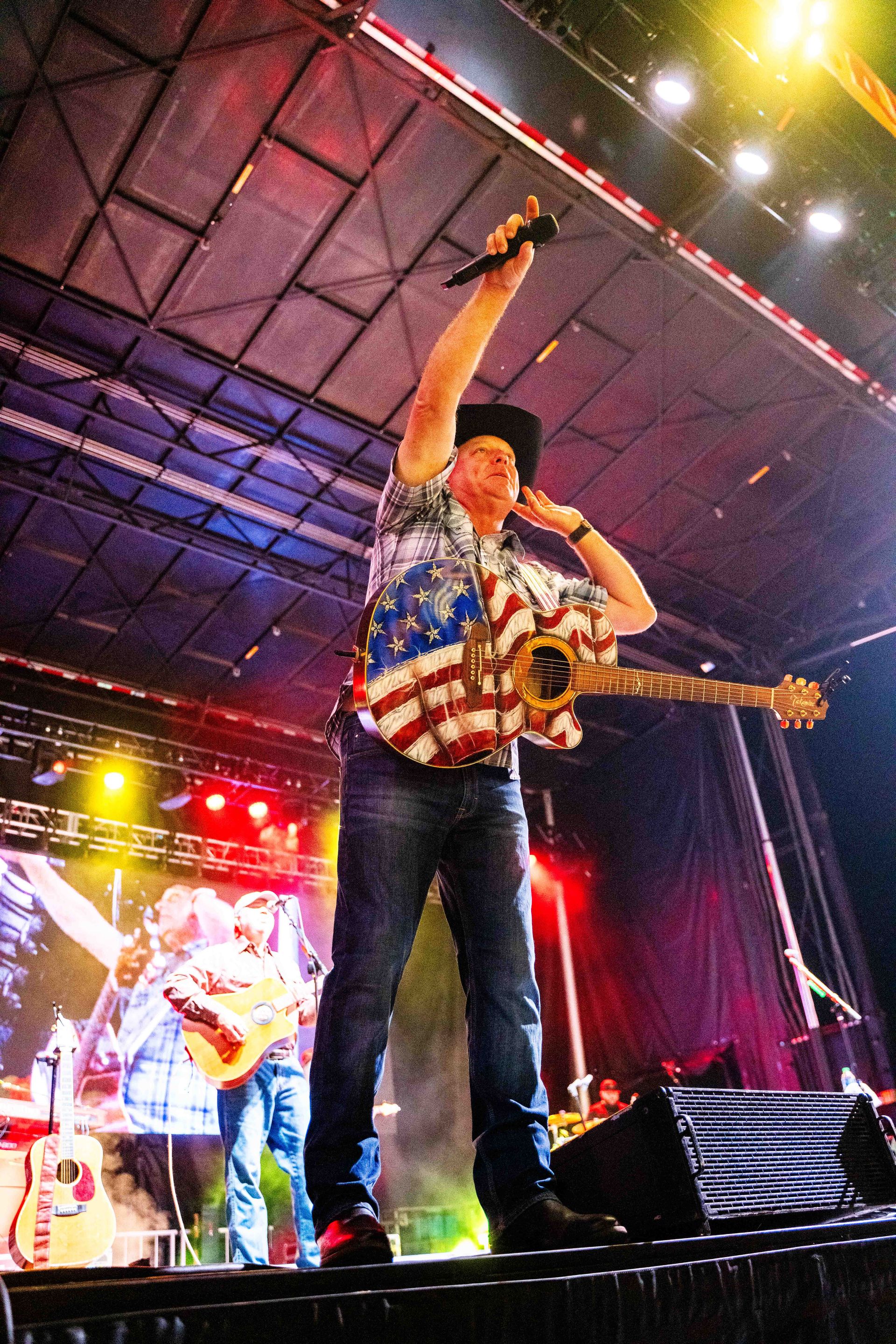 A man is singing into a microphone on a stage while holding an american flag.