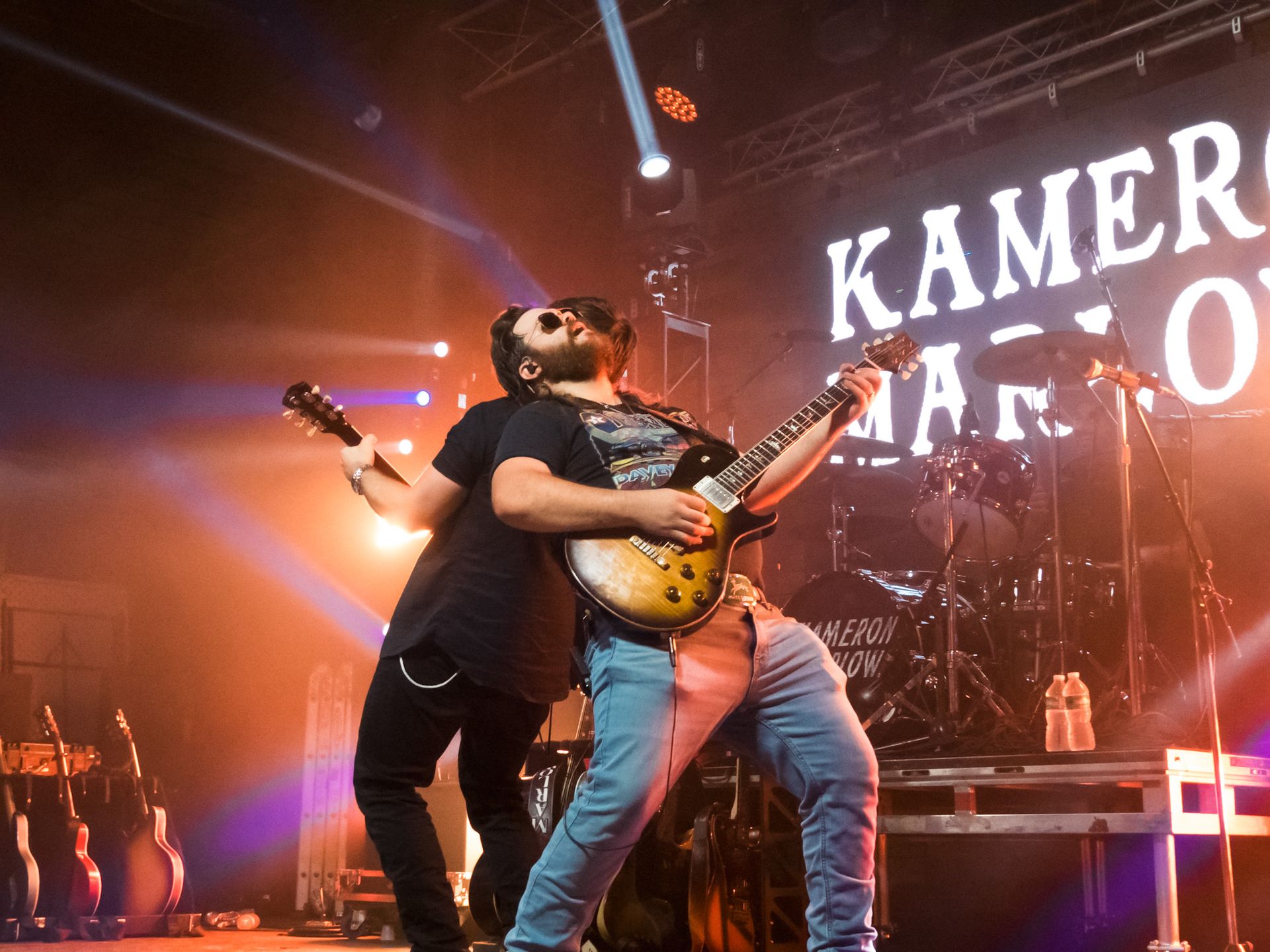 Two men are playing guitars on a stage in front of a sign that says kamero