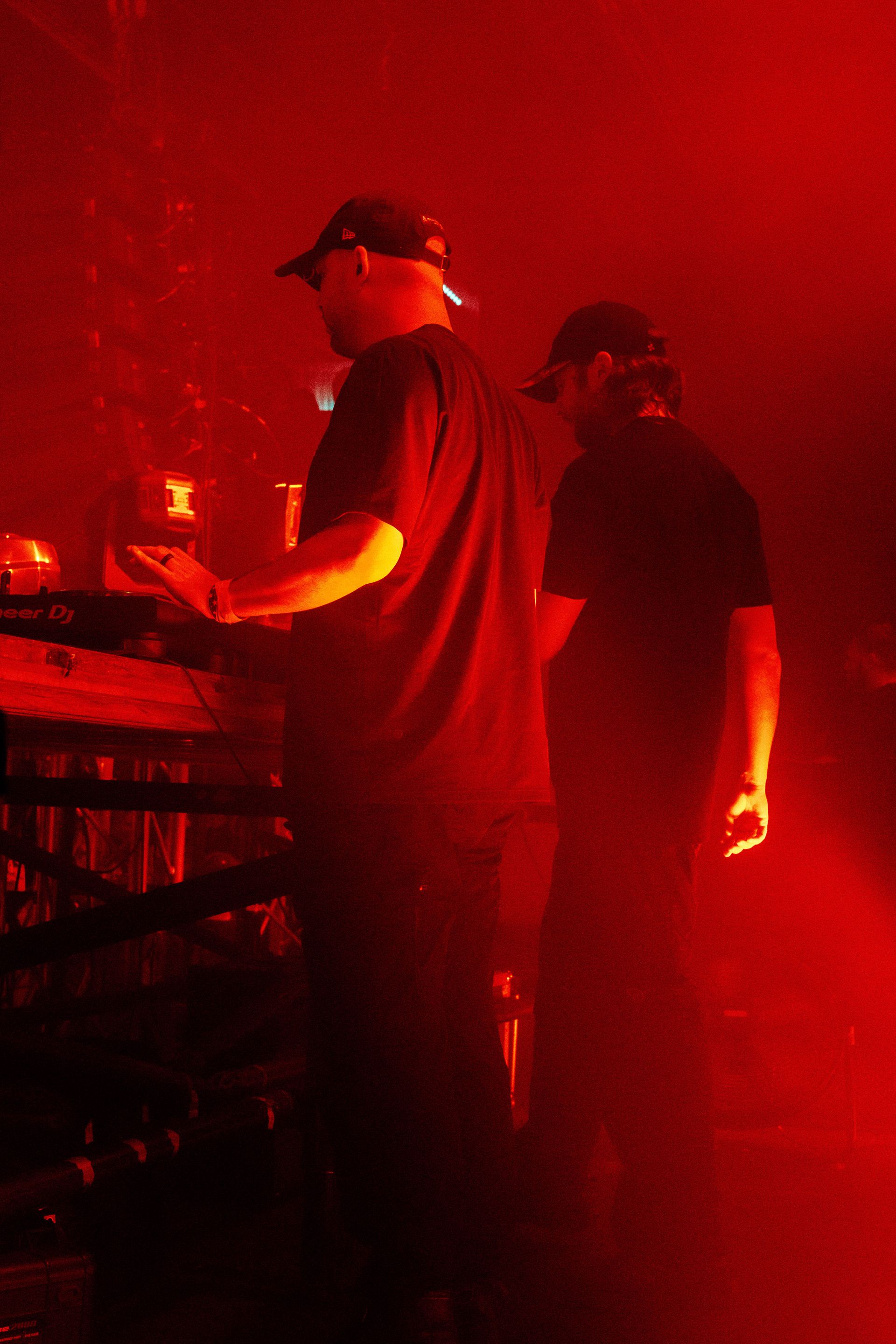 Two men are standing in a dark room with red lights behind them