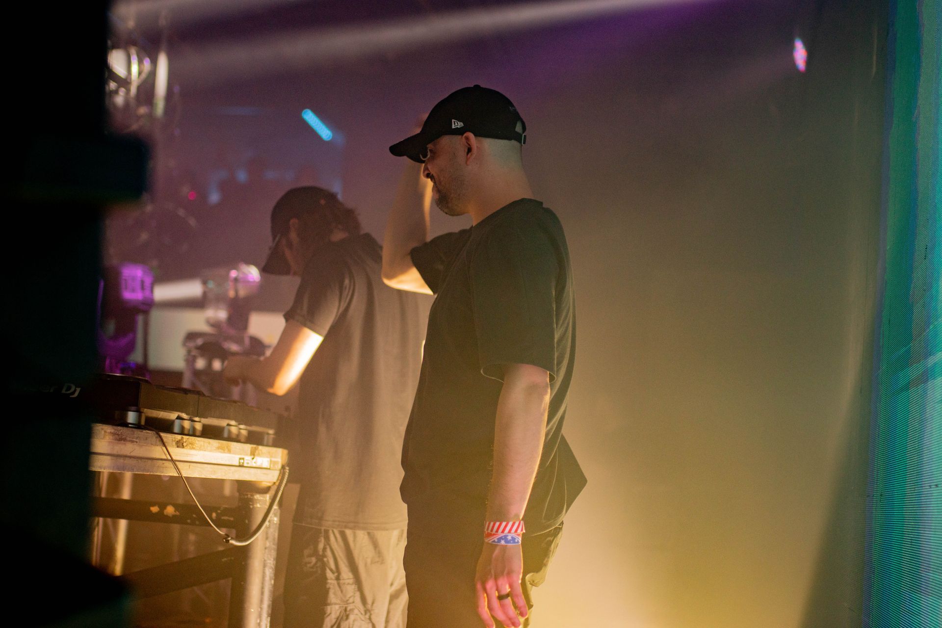 A man in a hat is standing next to another man in a dark room.