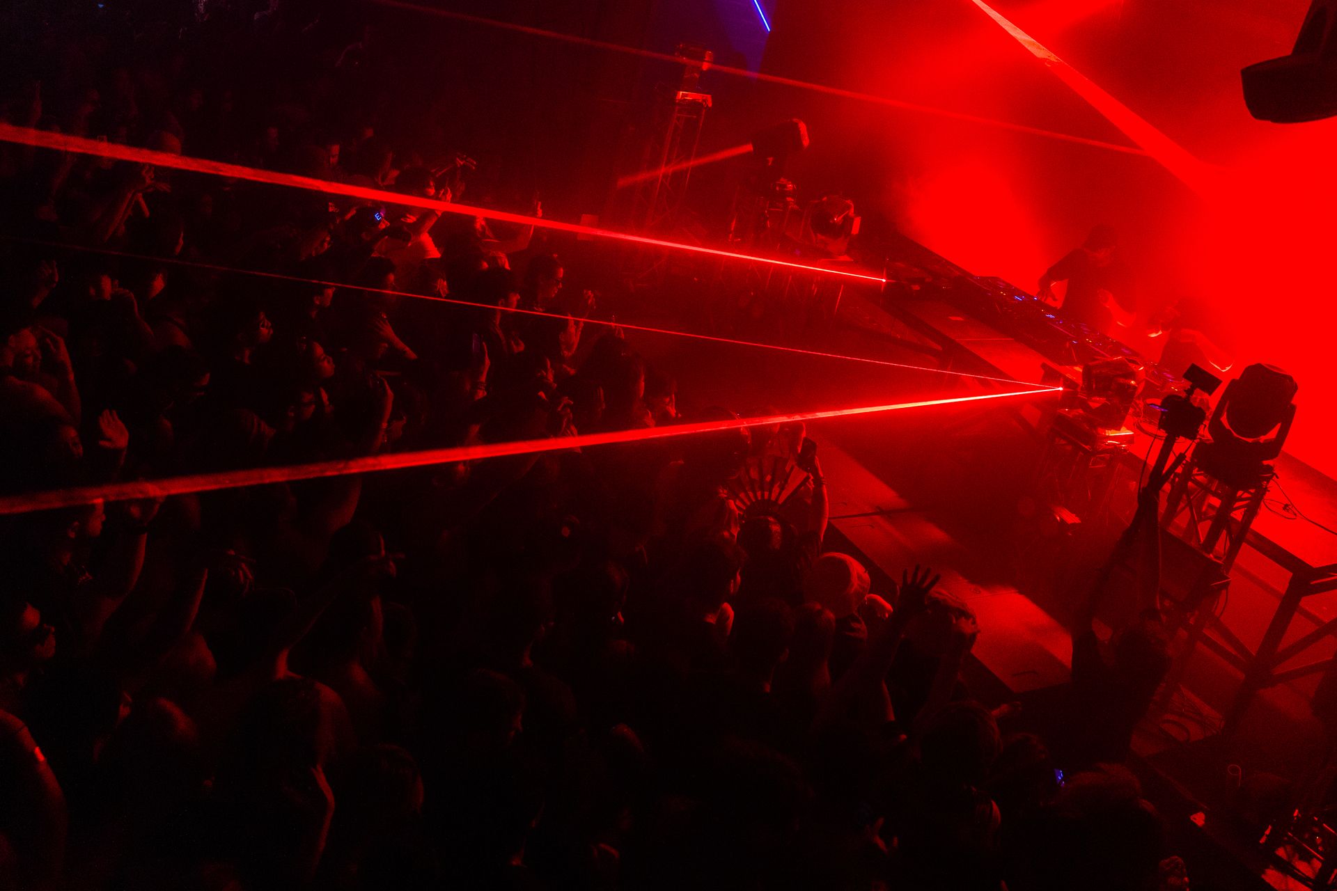 A crowd of people are watching a concert in a dark room with red lights coming out of the ceiling.
