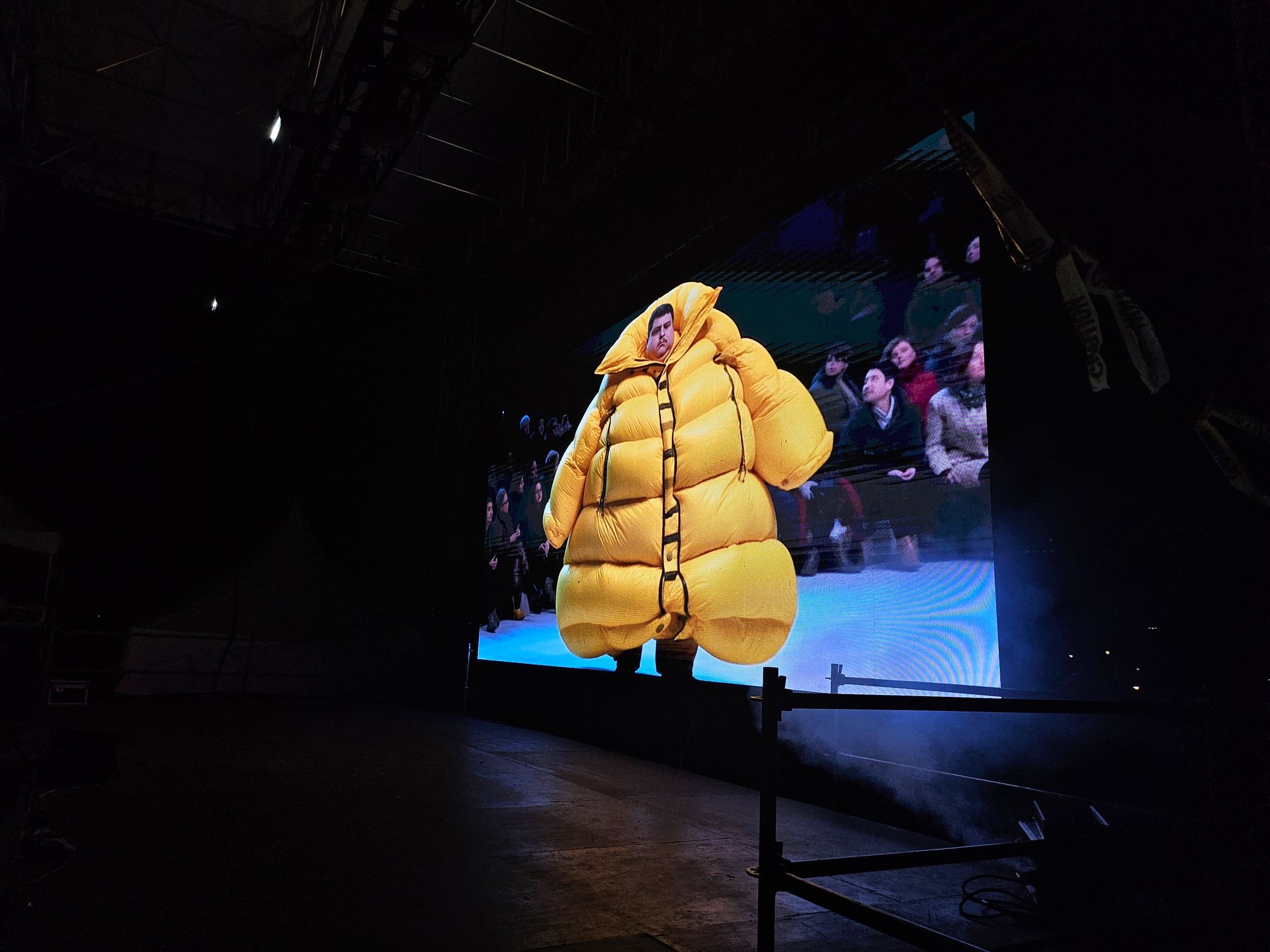 A person in a yellow inflatable suit is standing in front of a large screen.