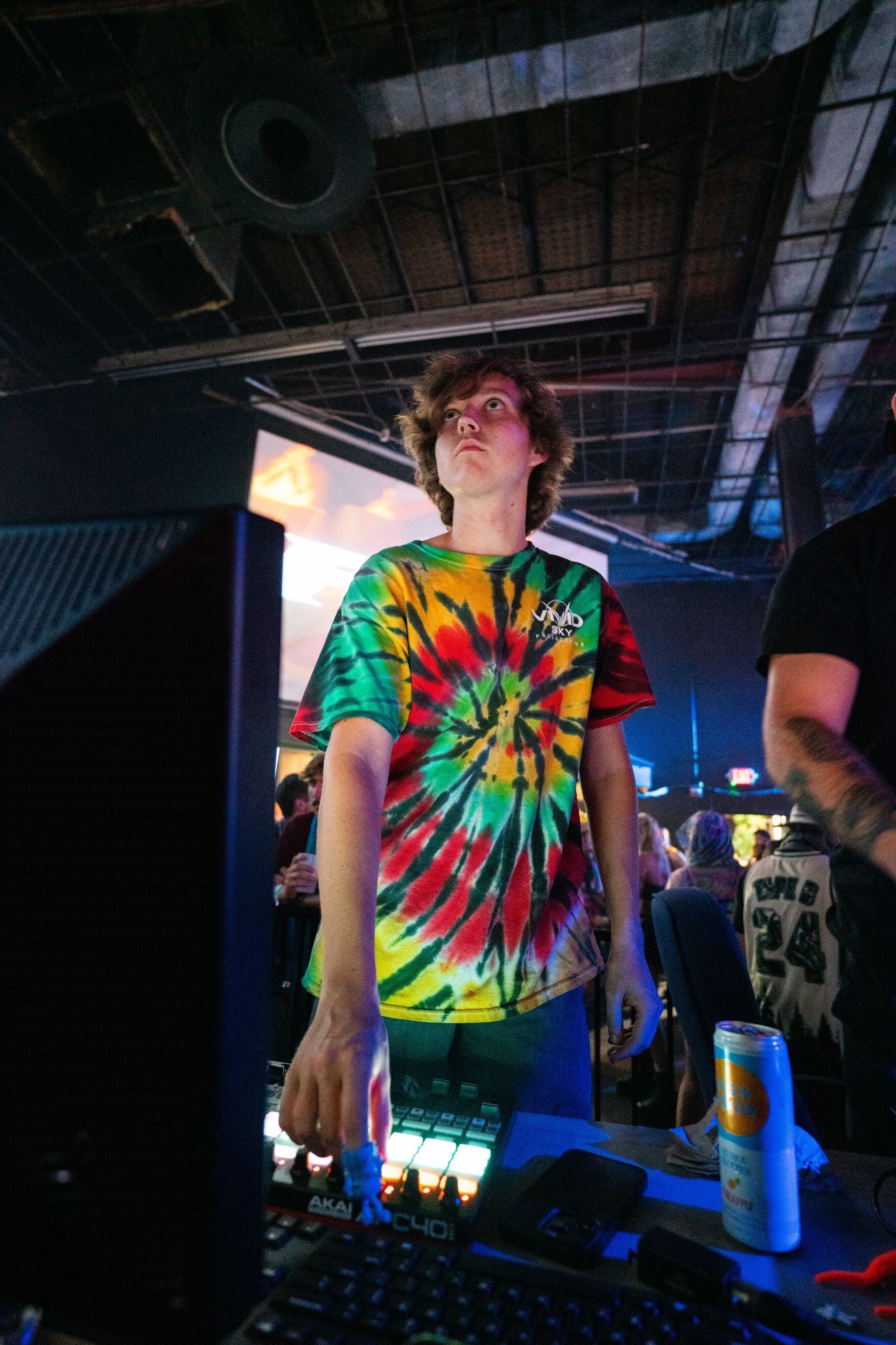 A man in a tie dye shirt is standing in front of a computer in a dark room.
