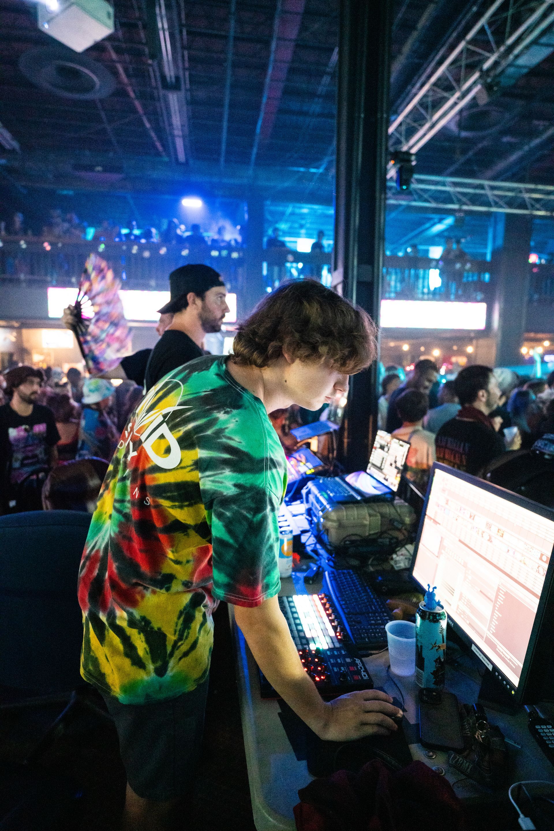 A man in a tie dye shirt is standing in front of a computer in a crowded room.