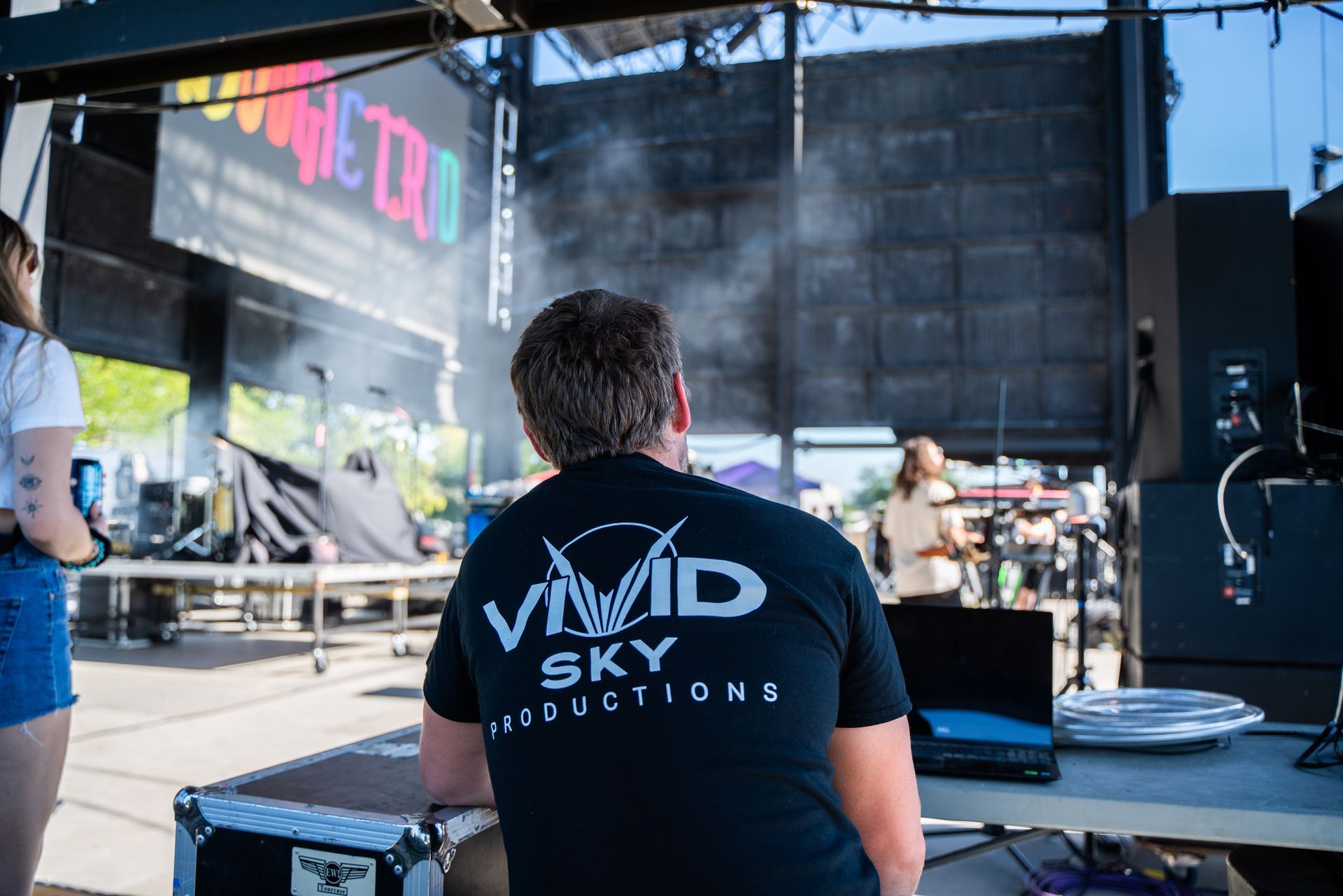 A man in a vivid sky productions shirt is sitting in front of a large screen.