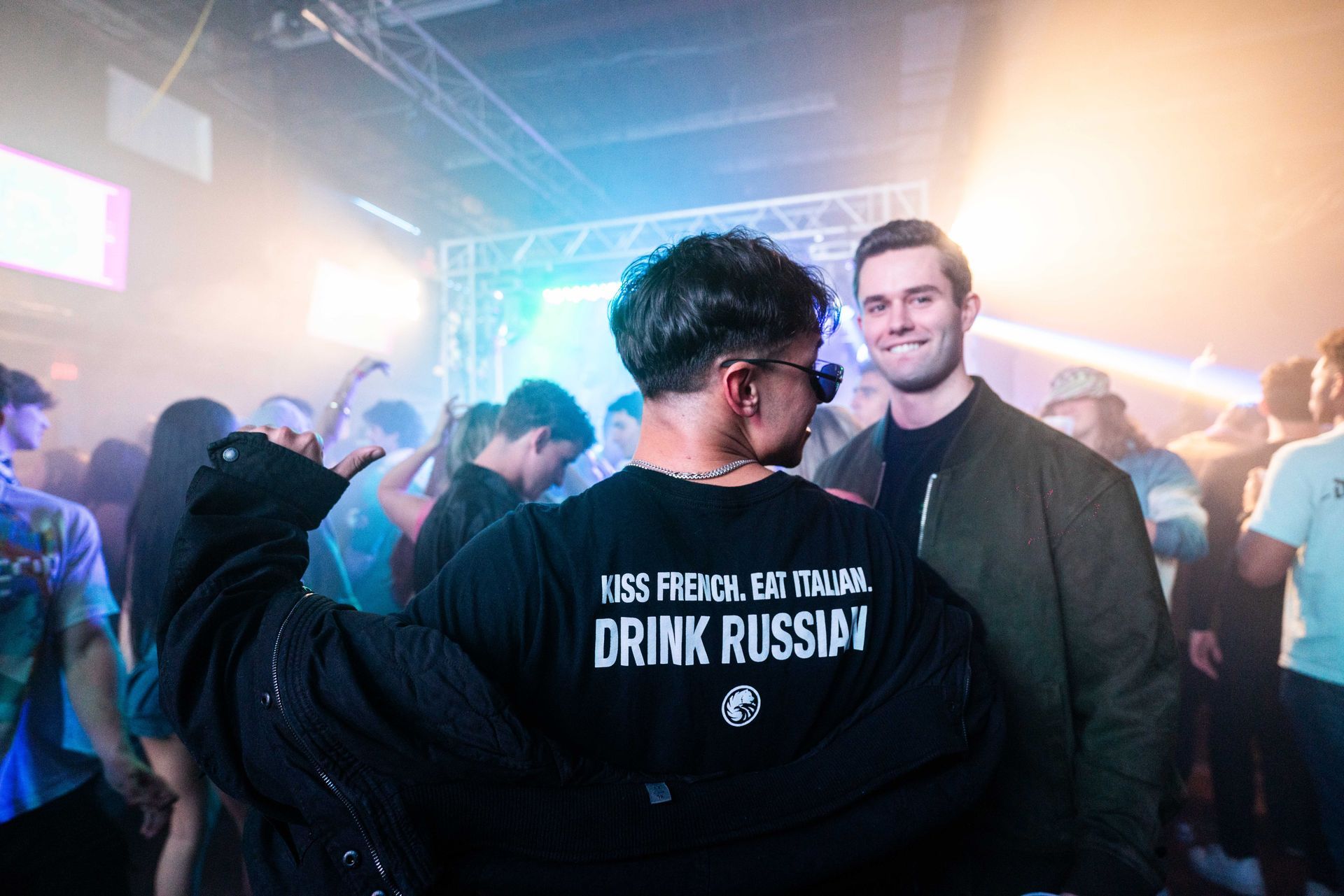 A man wearing a drink russian t-shirt is standing in a crowd of people at a party.