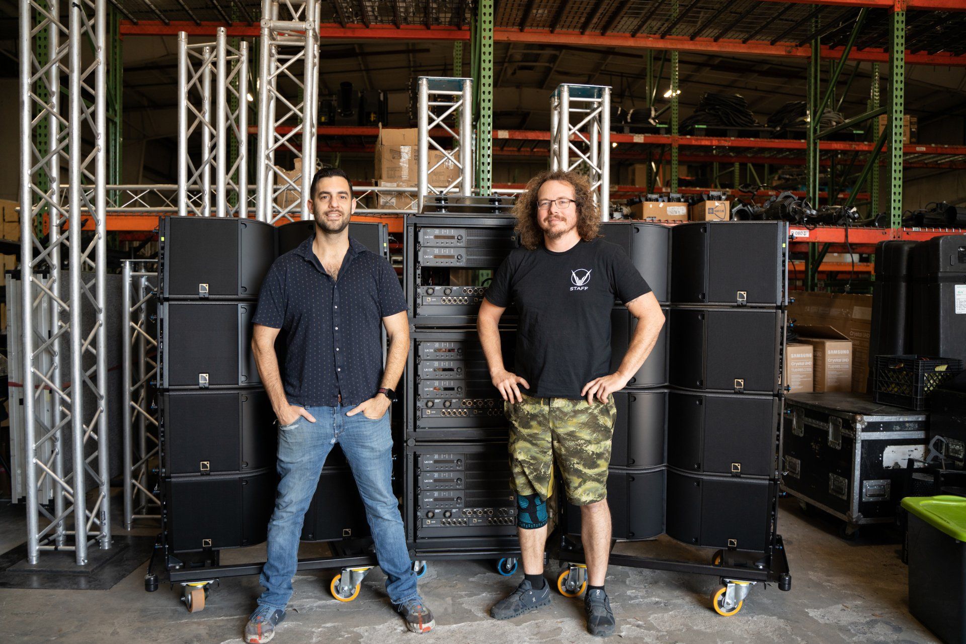 Two men are standing in front of a row of speakers in a warehouse.