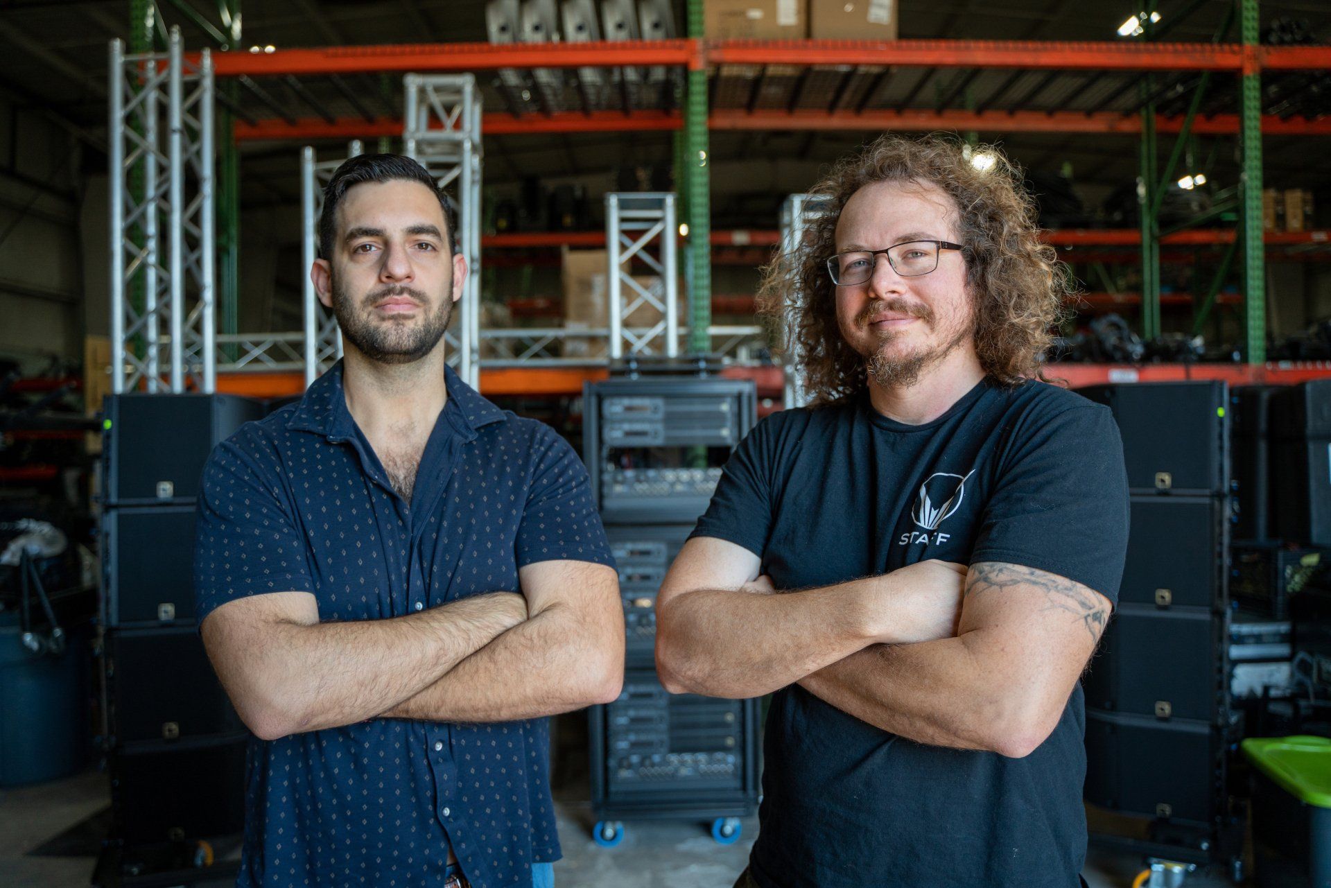 Two men are standing next to each other in a warehouse with their arms crossed.