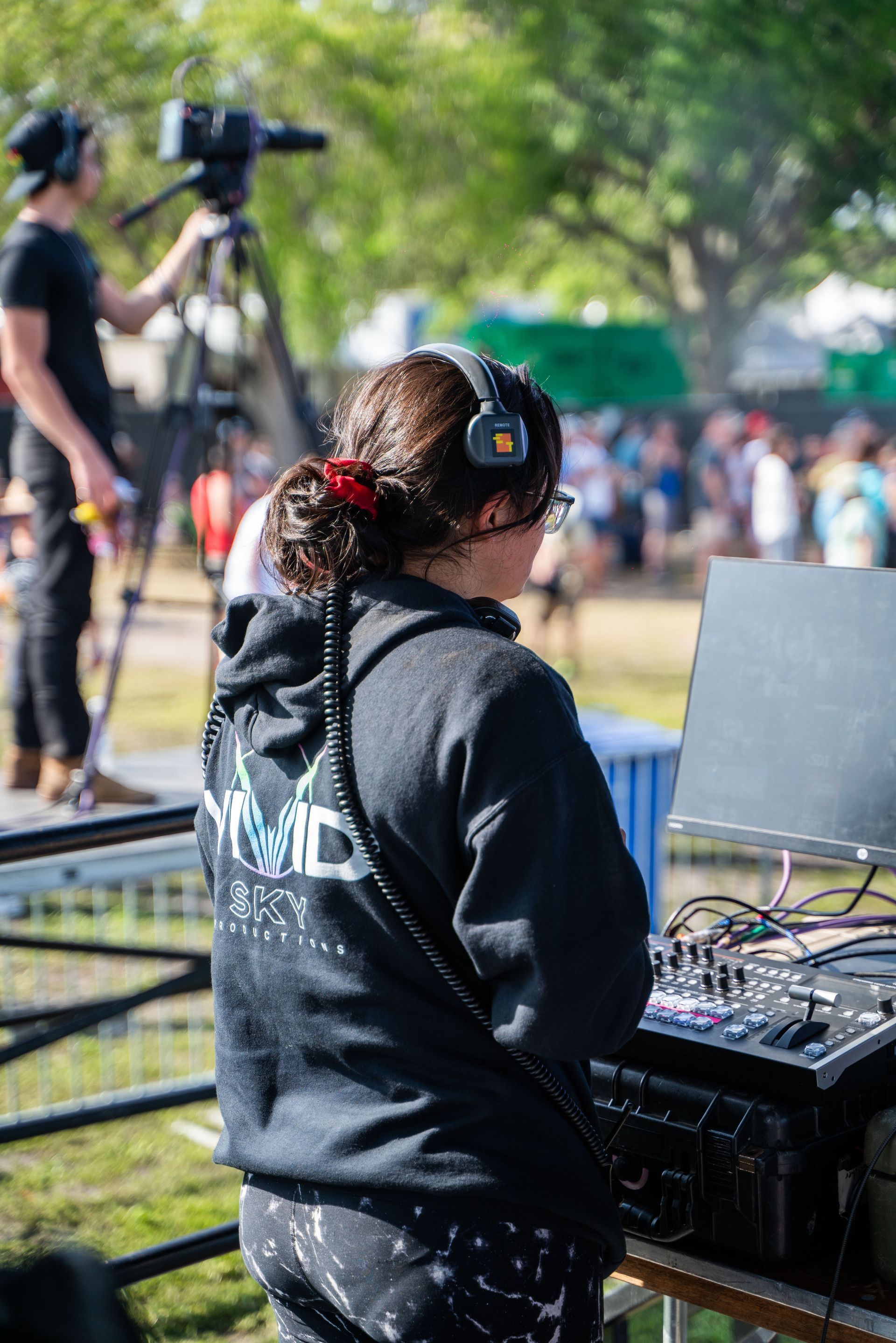 A woman wearing headphones is standing in front of a computer and a camera.