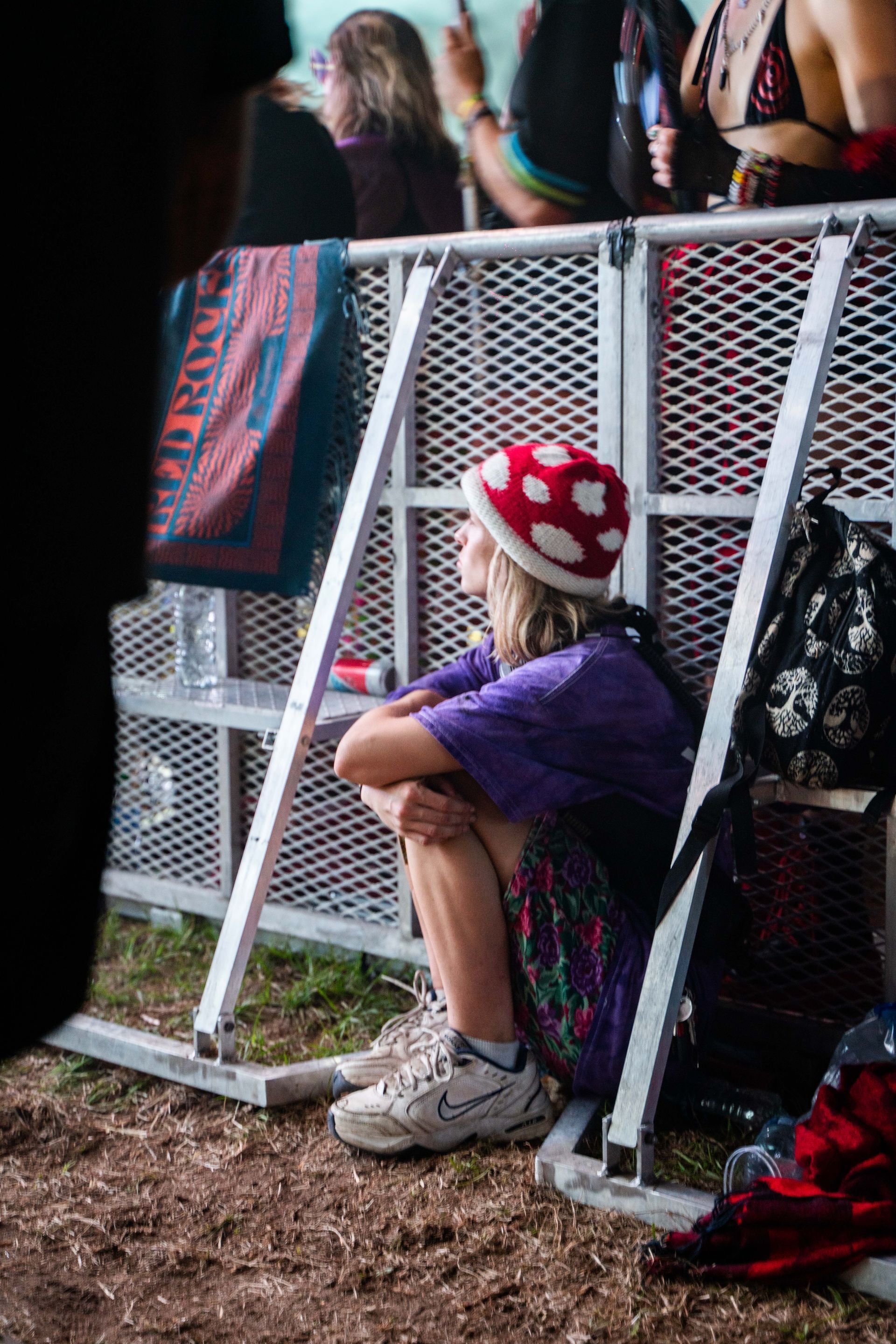A woman is sitting on the ground in front of a metal fence at a music festival.