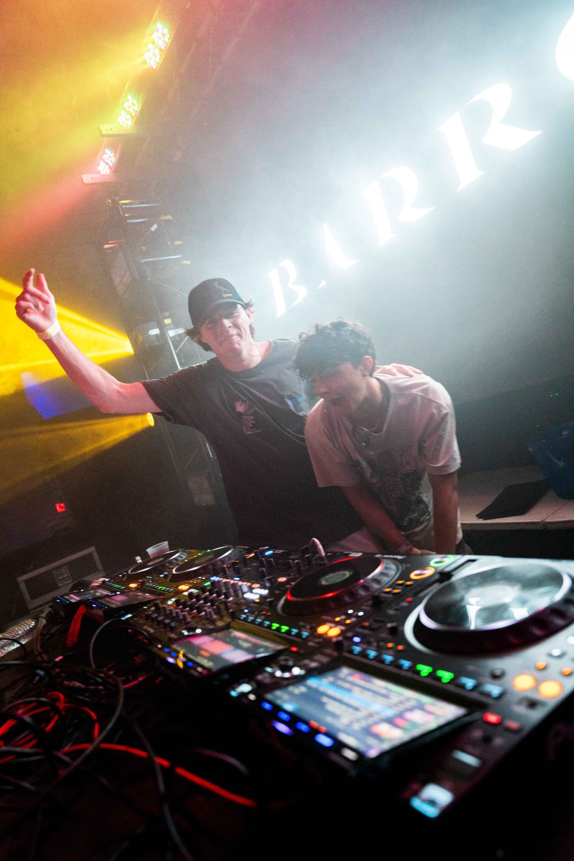 Two men are standing next to each other in front of a dj mixer in a dark room.