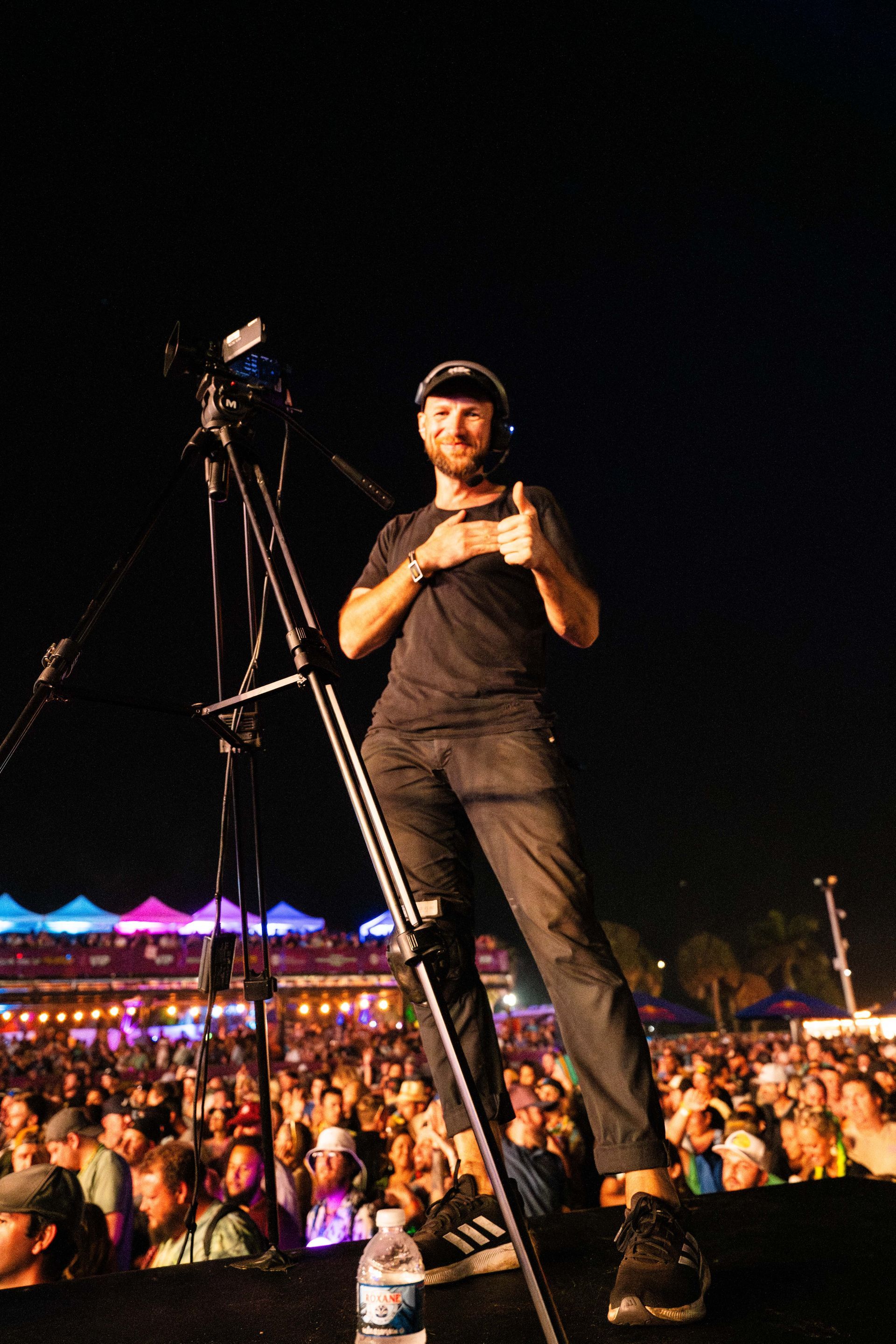 A man is standing on a tripod in front of a crowd and giving a thumbs up.