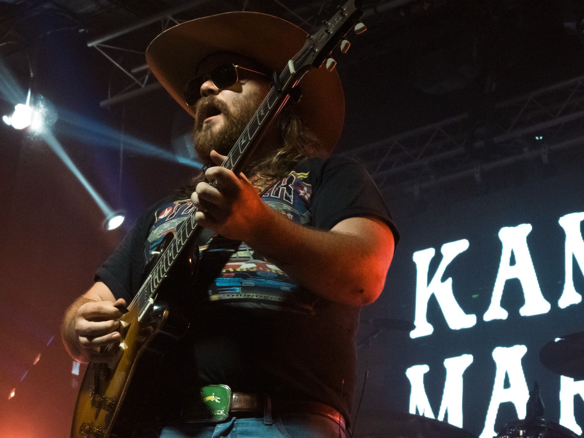 A man in a cowboy hat is playing a guitar in front of a sign that says kan man