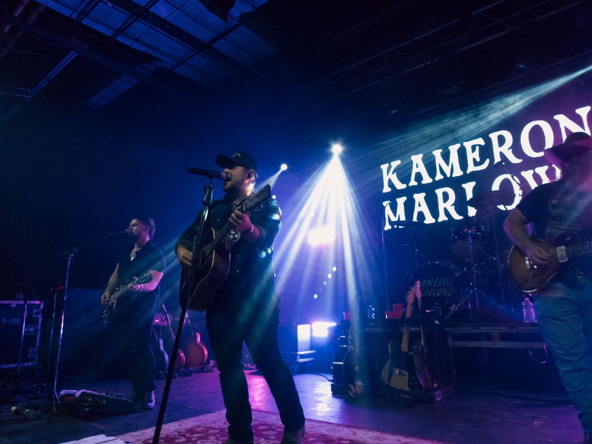 A man is singing into a microphone on a stage in front of a sign that says kameron mar !