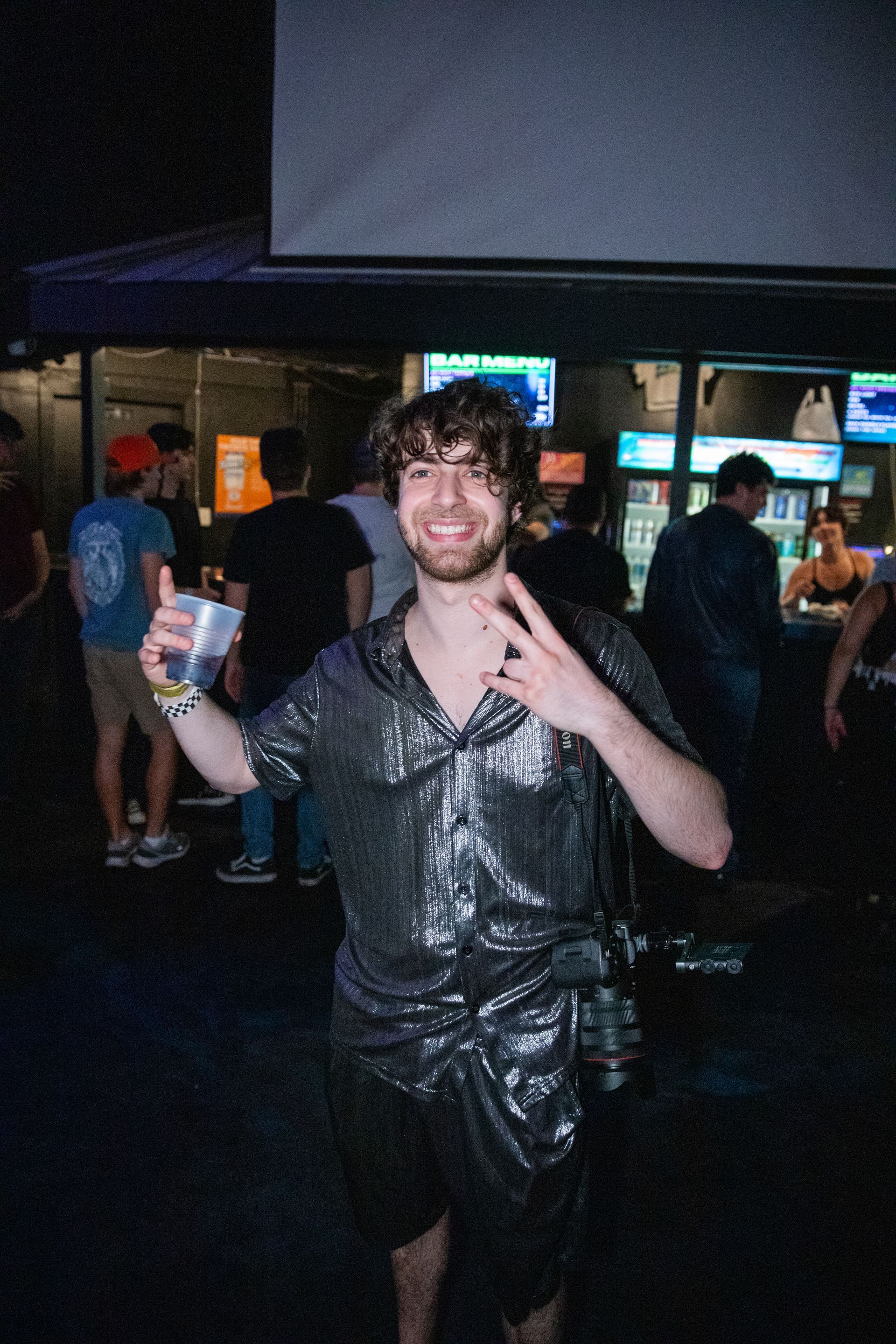 A man is standing in a crowd holding a cup and giving a peace sign.