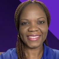 A smiling person with braided hair against a purple background, wearing a blue shirt.