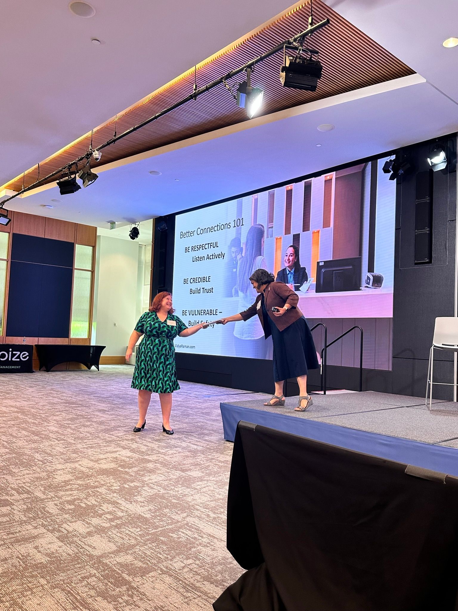 Two people shake hands on a stage in a large conference room with a presentation screen in the background.