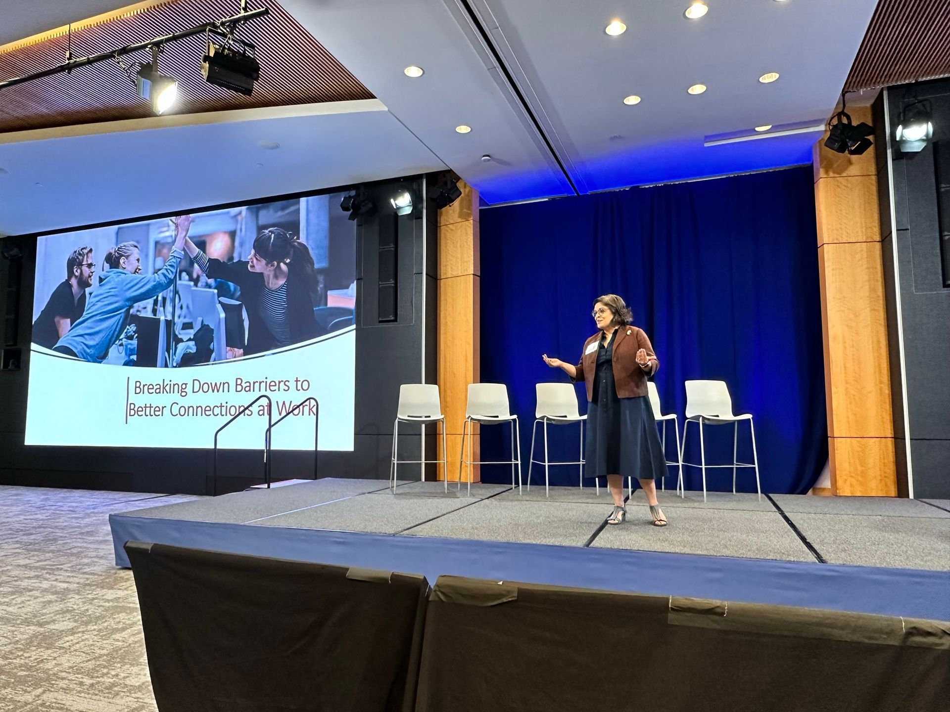 A person stands on a stage presenting in front of a screen displaying a photo of people high-fiving in an office setting.