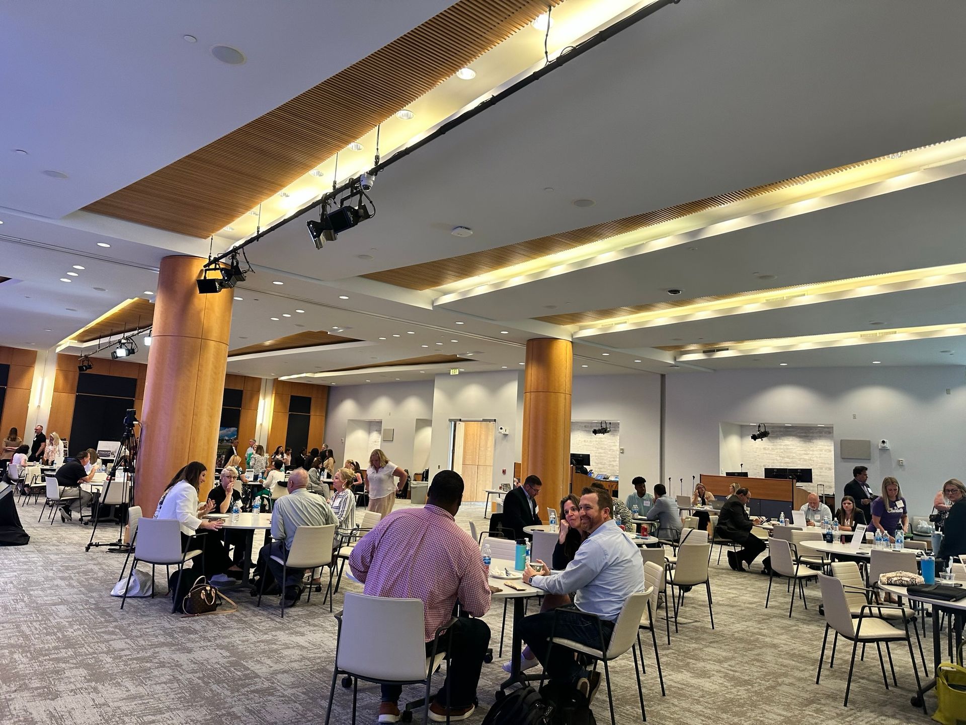 People seated at tables in a brightly lit, modern conference hall with wood-accented pillars and track lighting.