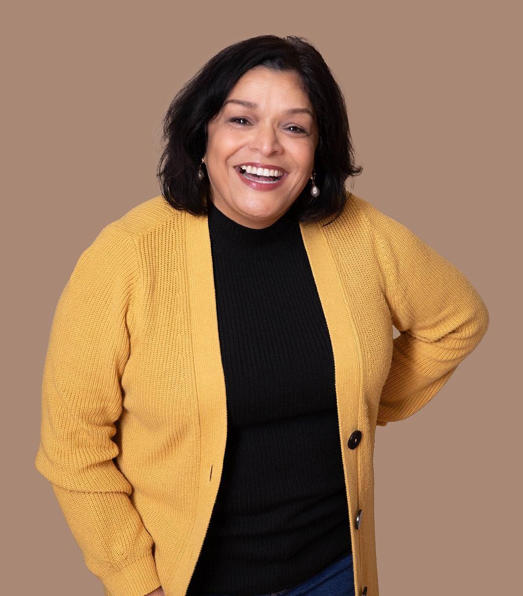Smiling person wearing a yellow knit cardigan over a black top, posed against a solid tan background.
