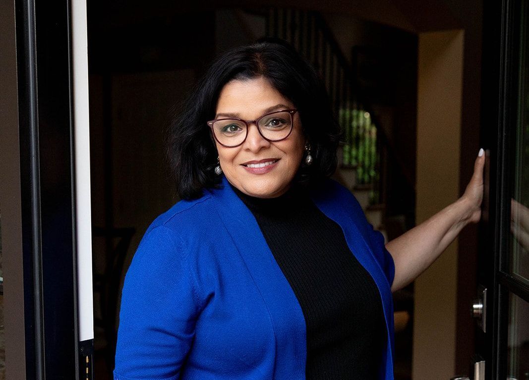 A person with dark hair and glasses wears a blue cardigan and black shirt while leaning against a doorway.