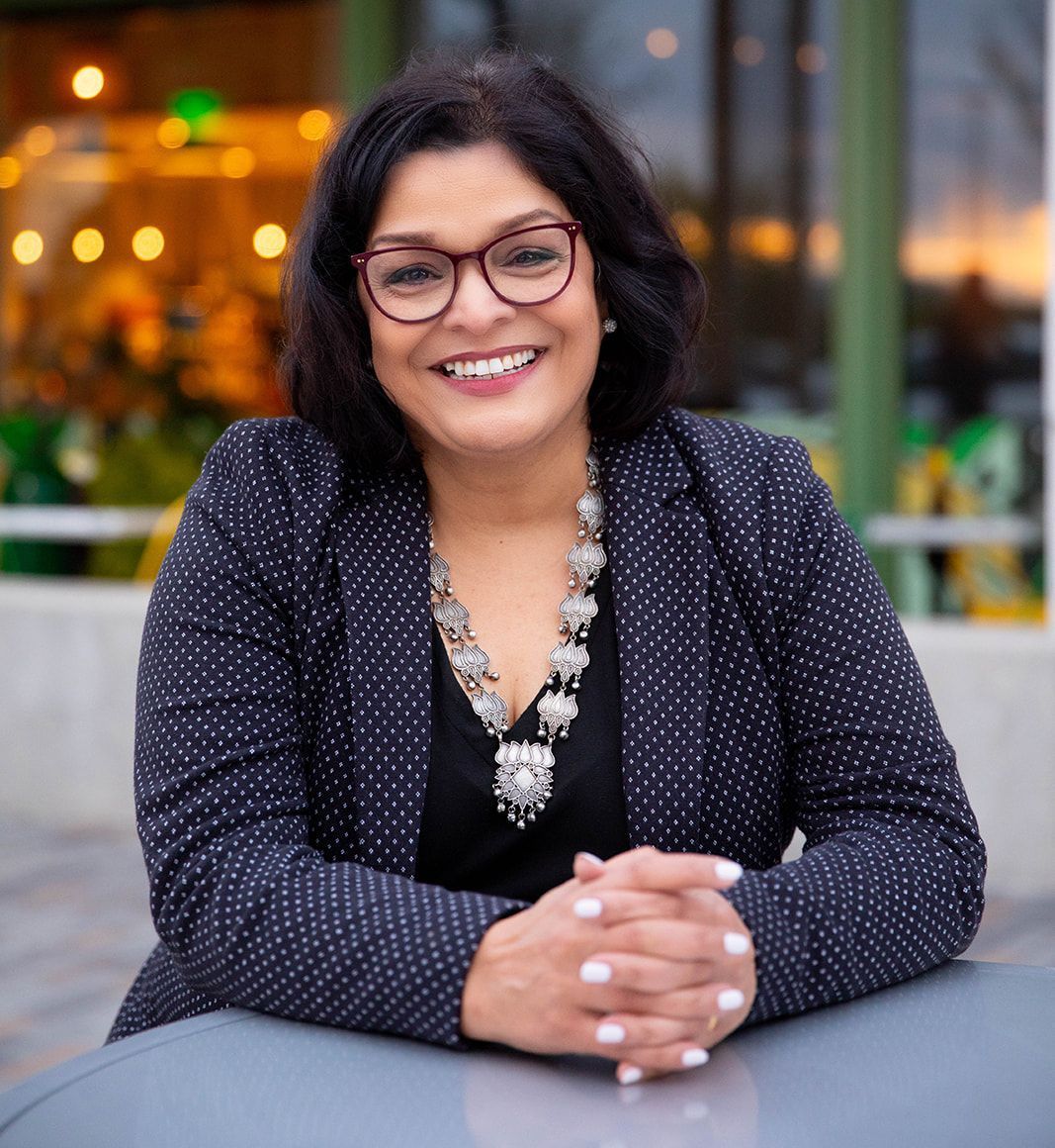 A smiling person with dark hair and glasses, wearing a dark patterned blazer and statement necklace, resting on a table.