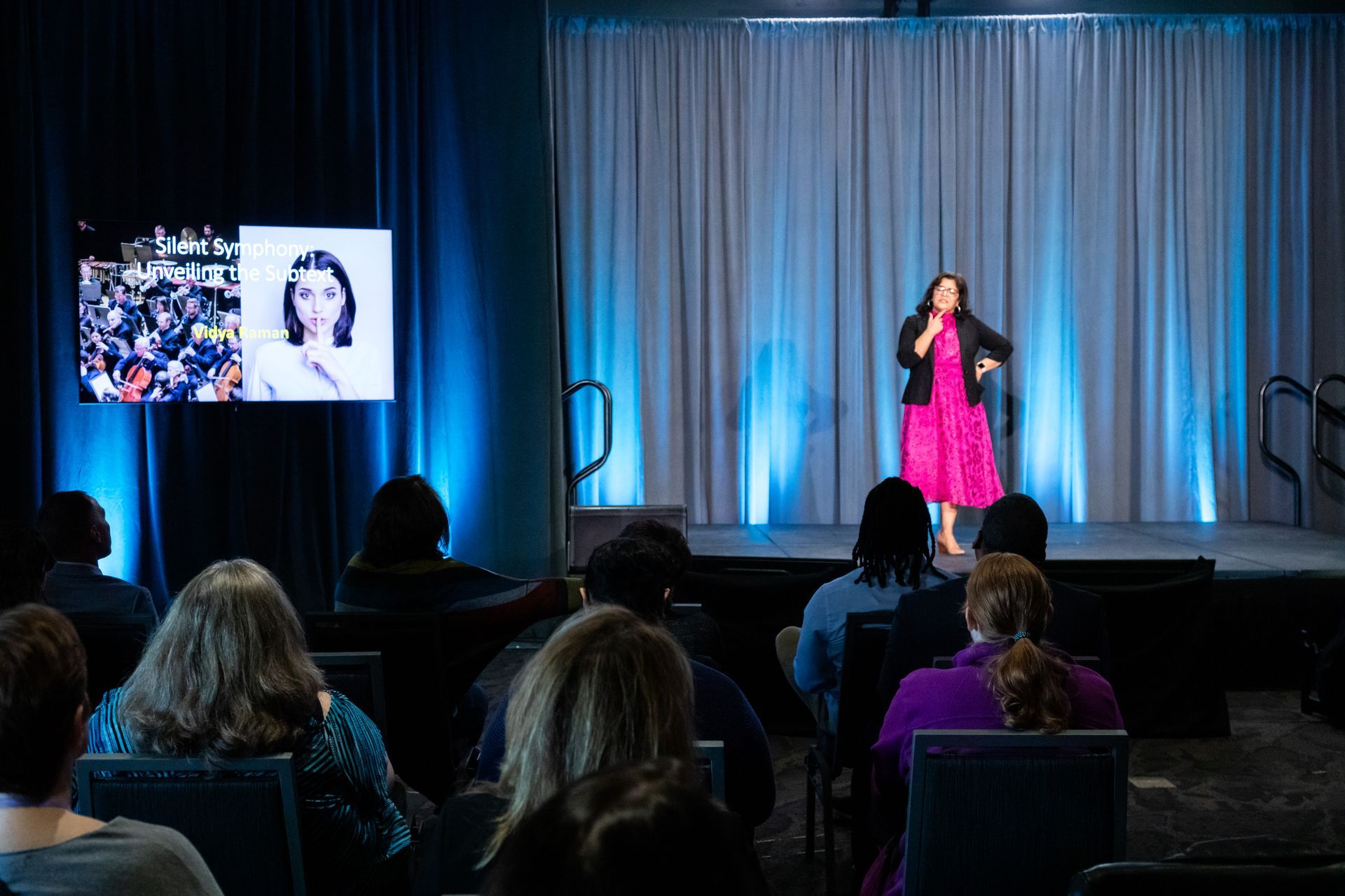 A speaker in a bright pink dress presents on stage to an audience, with a large screen displaying two photos beside her.