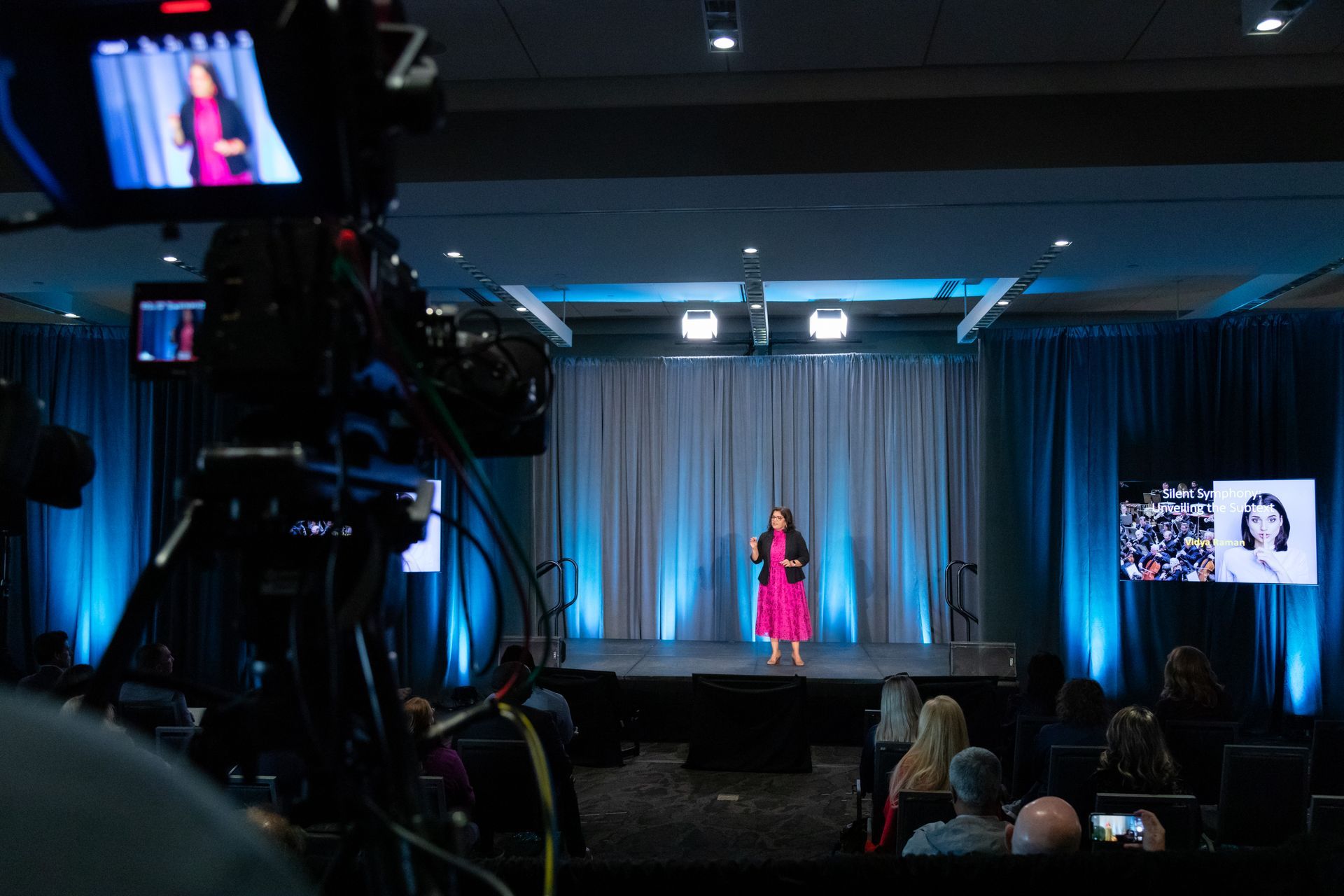 A person in a pink dress speaks on a stage with blue curtains before an audience, viewed past a professional video camera.