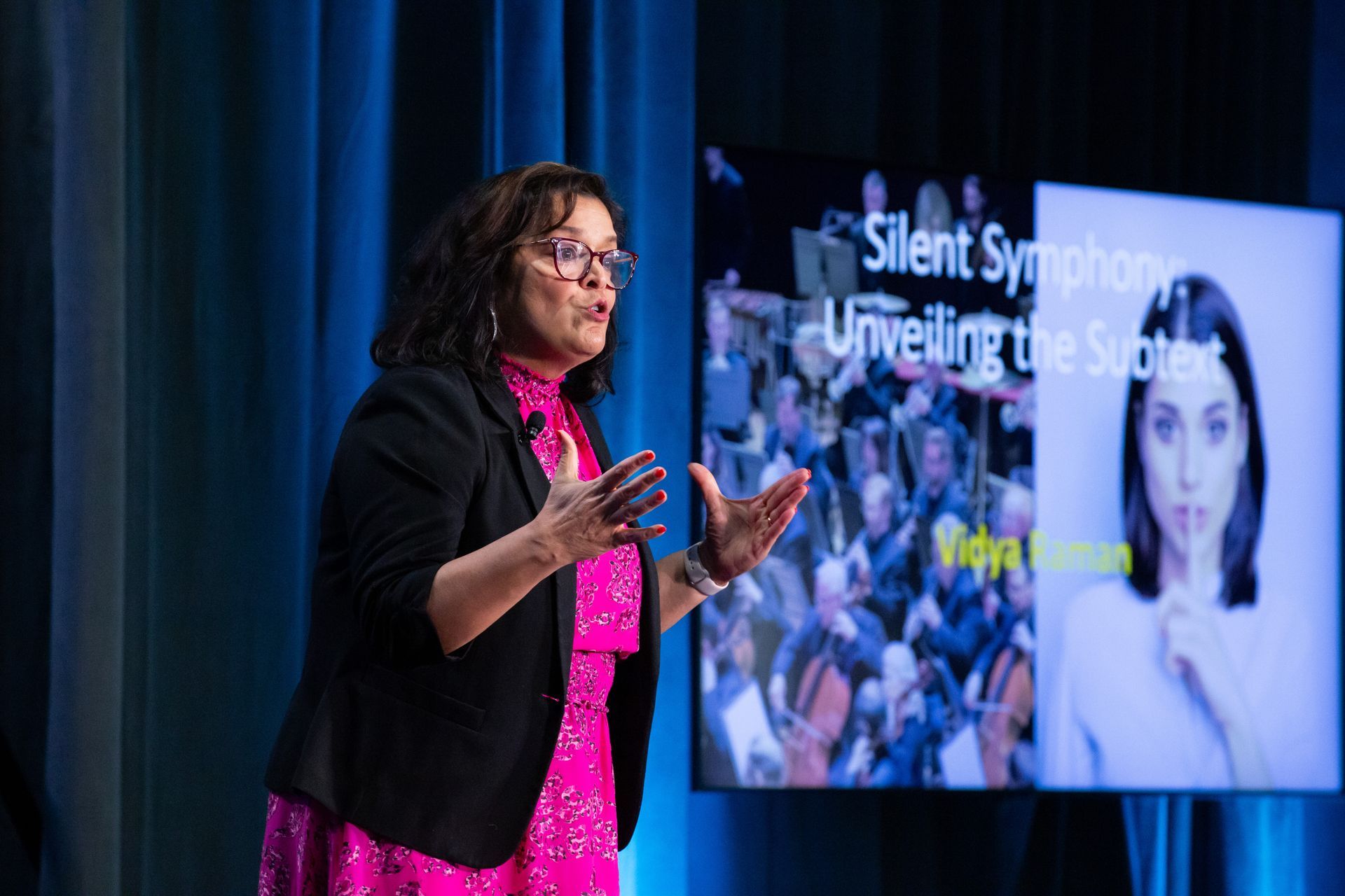 A speaker in a bright pink dress and black blazer gestures while presenting in front of a screen titled