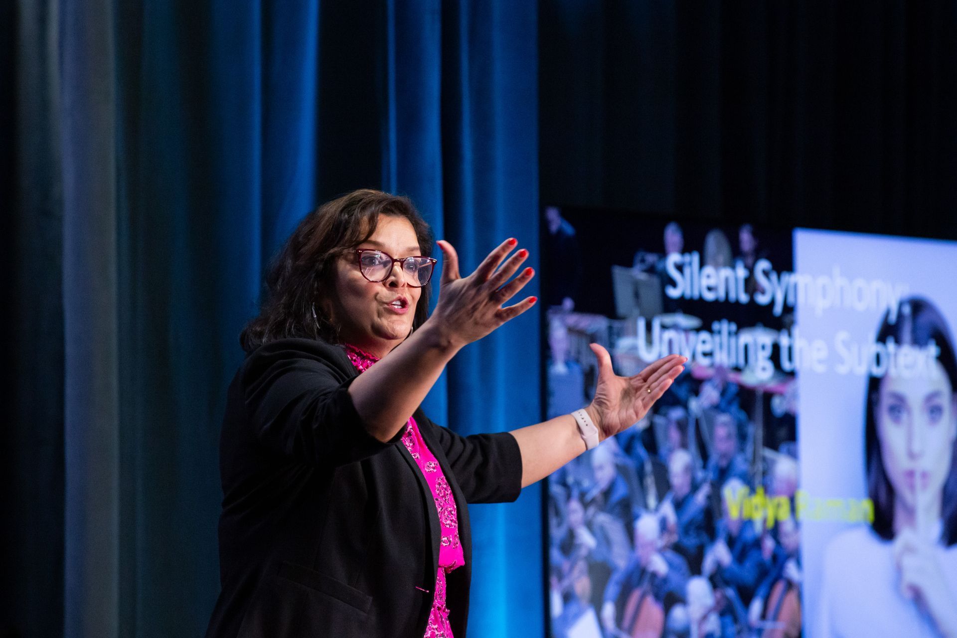 A speaker gesturing on stage in front of a screen titled