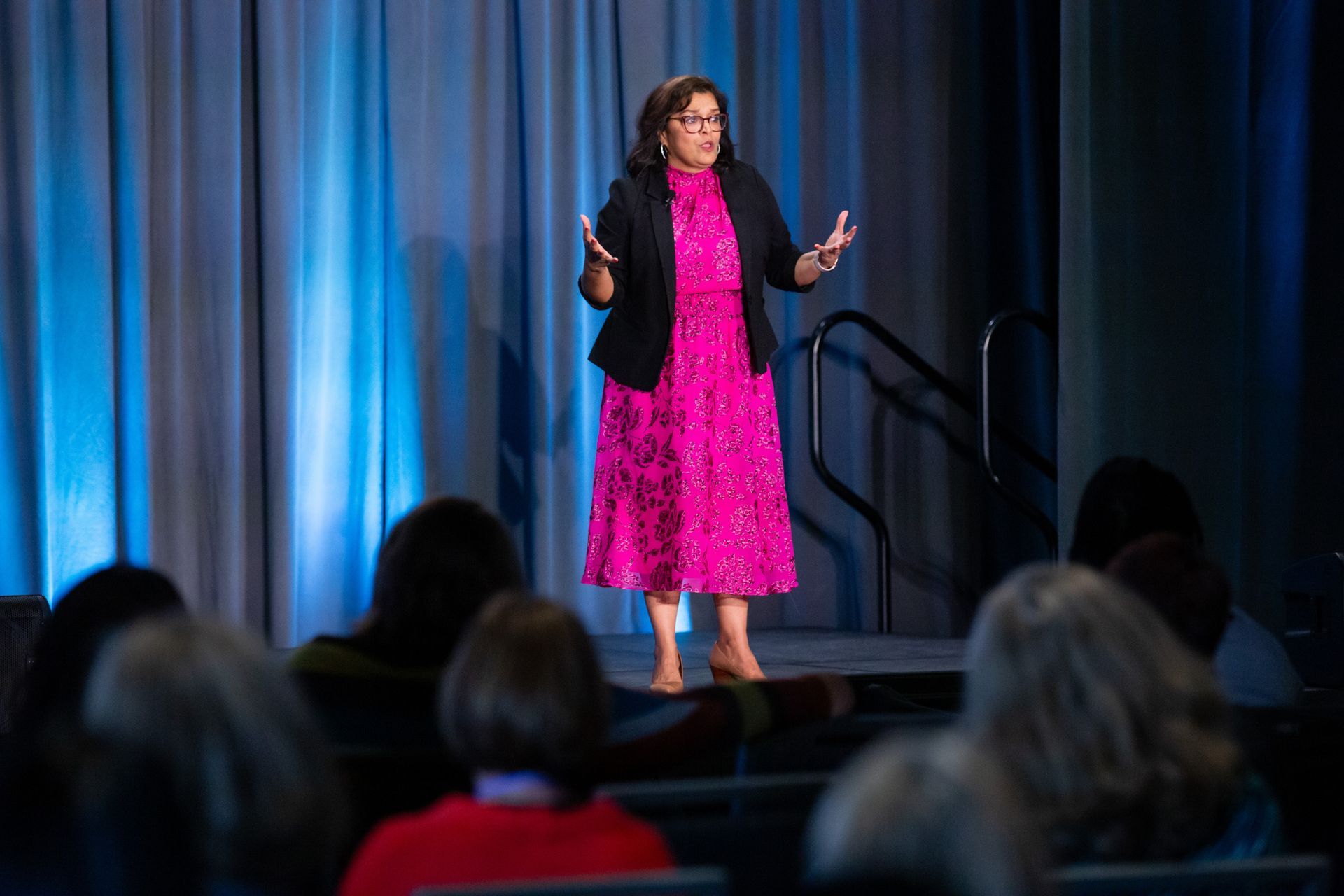 A person in a bright pink dress and black blazer stands on a stage gesturing while speaking to an audience.