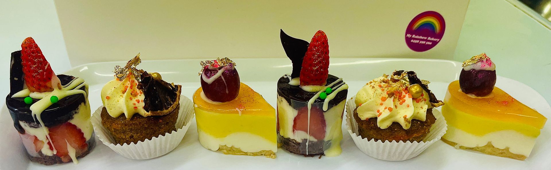A row of six miniature decorated pastries on a white plate, in front of a box — My Rainbow Bakery, Cafe & Catering in Cannonvale, QLD
