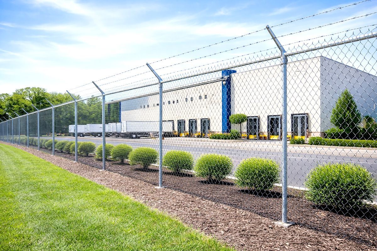 Chain link fence with barbed wire around a warehouse. Green grass and bushes in front.