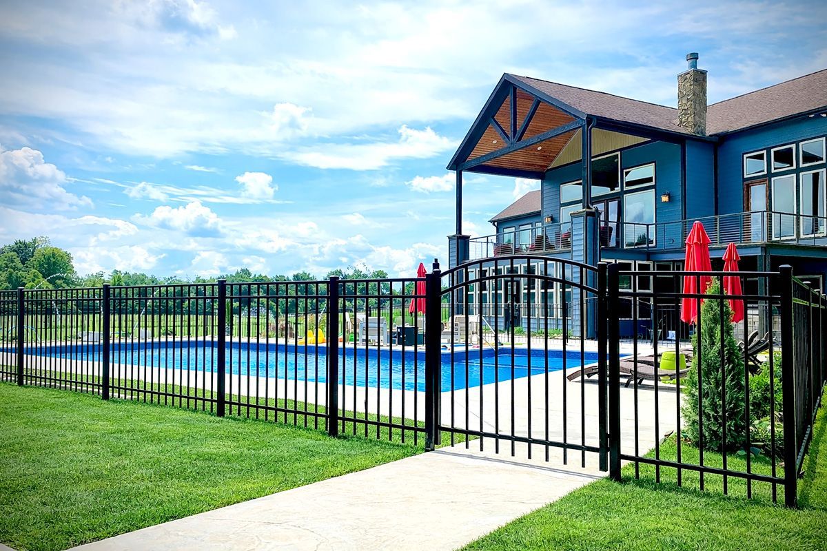 Black fenced-in swimming pool in front of a blue house with a covered porch under a blue sky.