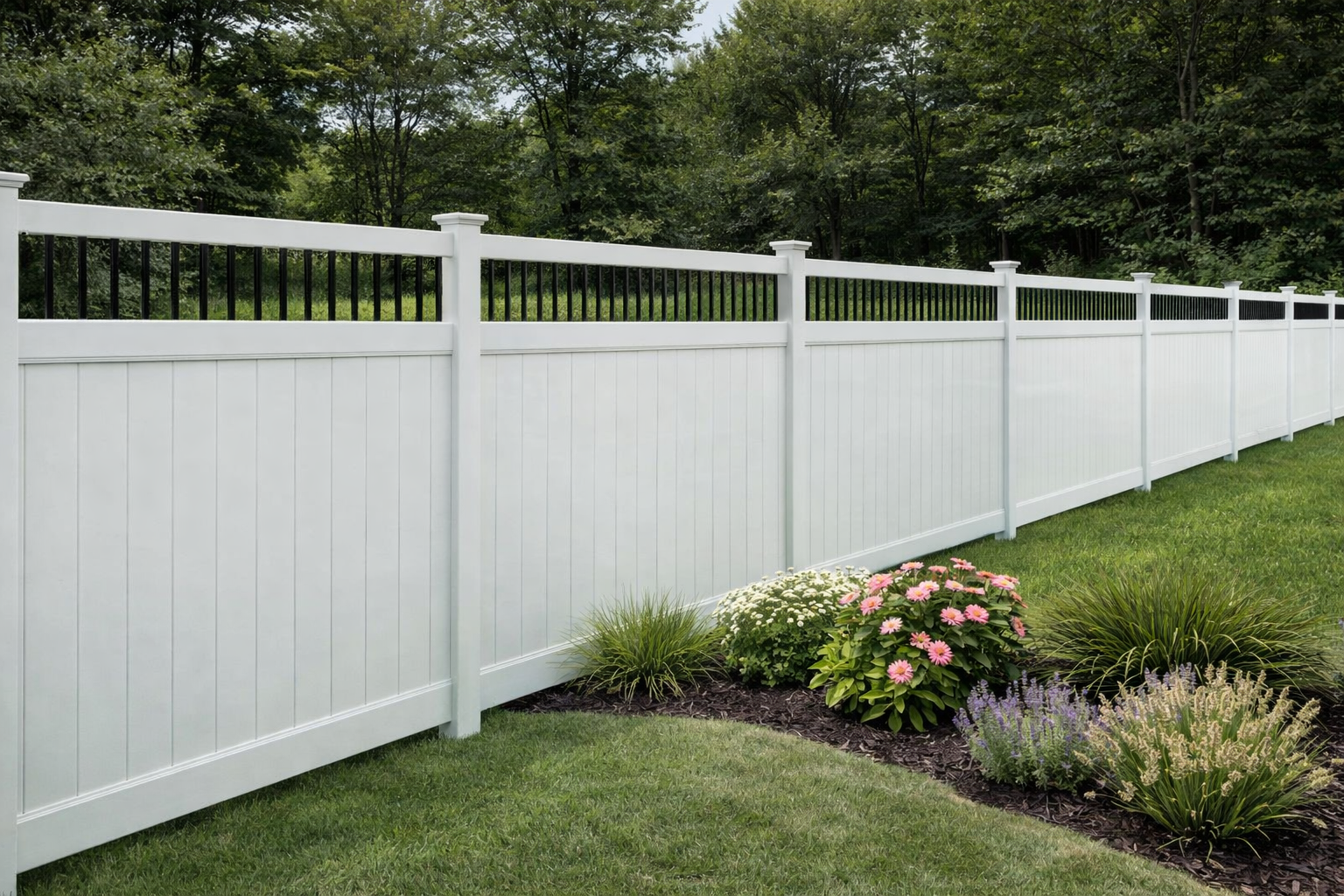 White vinyl privacy fence with black decorative top, in a grassy yard, with flower bed and trees.